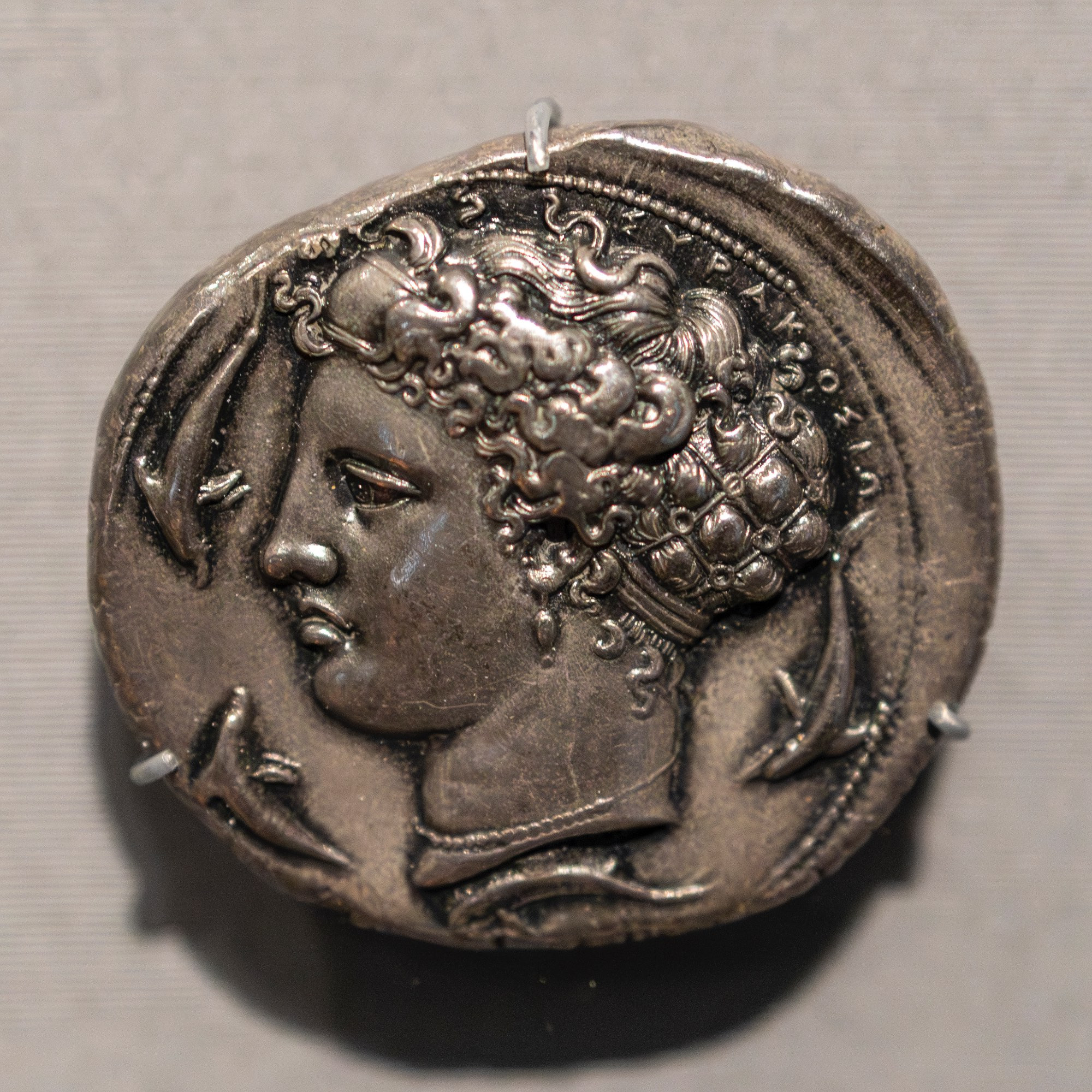 The reverse of this silver decadrachm depicts the profile of Arethusa, a nymph (or nereid) who was transformed into a freshwater spring on the tiny island of Ortygia adjacent to Syracuse, Sicily. 

As the patron figure of Syracuse, Arethusa is shown with her hair in a beautiful net (golden hairnets were a luxury of the elite) and surrounded by swimming dolphins. An inscription above and to the side of her head reads: ΣΥΡΑΚΟΣΙΩ[Ν], or Syrakosion - Syracuse. 

Greek, minted in Syracuse, Sicily, Italy (part of Magna Graecia, Great Greece - the Greek colonies in Italy), ca. 405-400 BCE. Silver.

Diameter: 37.8 mm (1.488 in.)
Weight: 43.11 g 

Getty Villa Museum, on loan from the American Numismatic Society (1964.79.21)