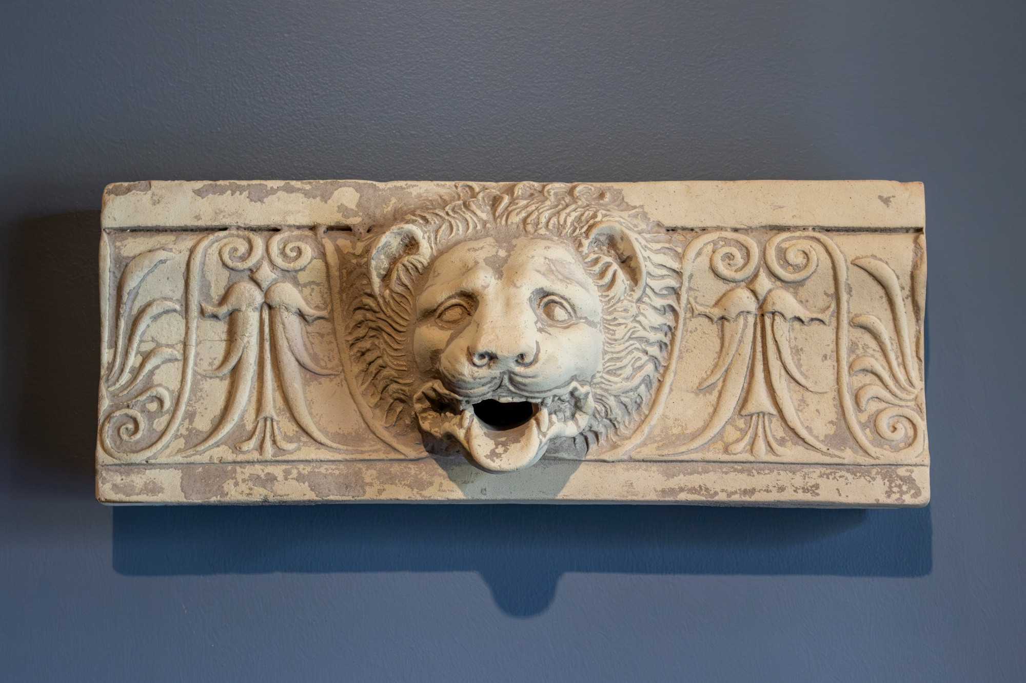 In the center of this sima (a projecting molding from the edge of a roof) is a waterspout fashioned in the shape of a lion's head. The lion’s mouth is open, revealing its teeth, and its tongue projects. The head is surrounded by a thick mane. On either side are inverted lotus and half-palmettes in relief. Some remains of pigments - particularly the white ground - can still be detected.

In classical architecture, a sima is the upturned edge of a roof which acts as a gutter. The term "sima" comes from the Greek simos, meaning bent upwards. A lateral sima runs along the horizontal edges and is broken by downspouts to let out rainwater.

Terracotta simas, like this one, have repeating patterns that are easy to reproduce with molds. They would have been brightly painted when new. Lion's heads were common, but ram and dog heads also existed. These animal heads may have served as religious symbolism, or as puns on the structure's geographic location.

Greek, South Italian, Metapontum, ca. 425-400 BCE. Terracotta with polychromy.

Getty Villa Museum (71.AD.449)