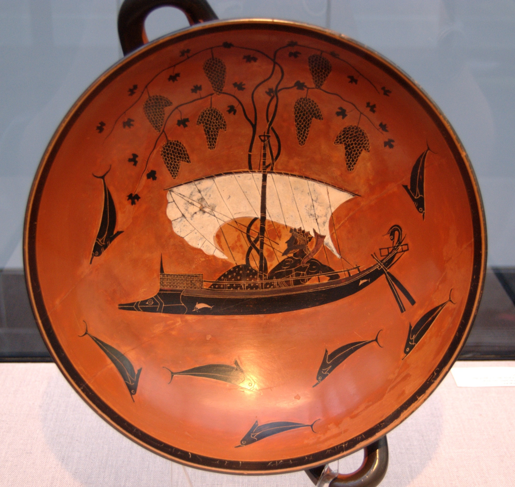One of the masterpieces of the Attic black-figure potter Exekias (Attic Greek), the inside of this kylix - a broad shallow drinking cup - depicts the god Dionysos, leaning back on a dark ship with a white sail. A large grape vine, heavy with bunches of fruit, has grown from the deck, and spreads out above the vessel. A multitude of dolphins - referencing the pirates transformed by Dionysos - leap in the waters about the ship.