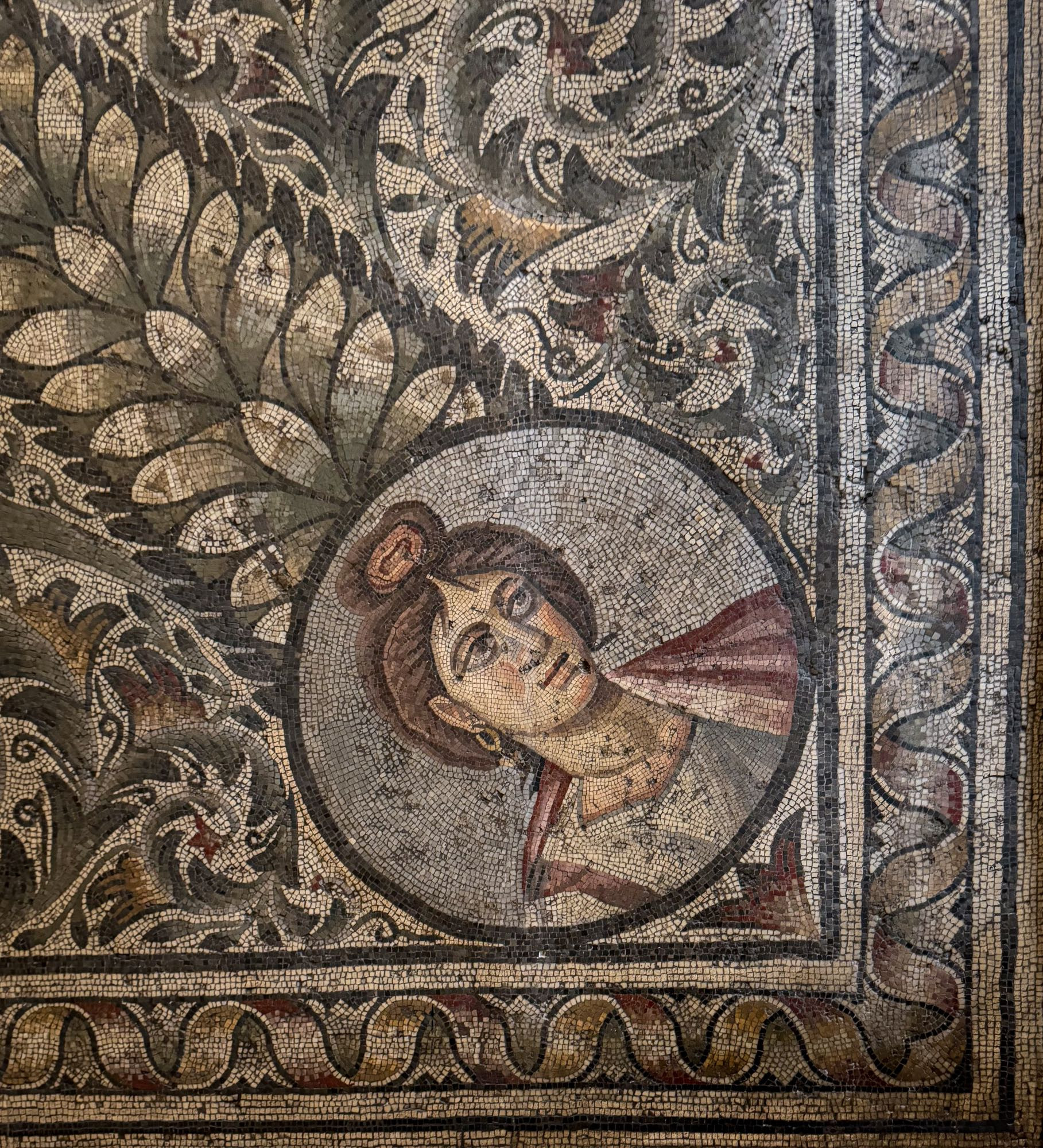 Corner of a large floor mosaic with a medallion in which is depicted a portrait bust of the female personification of Spring. She’s wearing a white tunic with a deep purple clavi (vertical stripe) and a reddish-purple mantle. Her hair is braided and pulled up into a topknot, as is often seen in Byzantine era art. She’s sporting simple gold hoop earrings. Surrounding this medallion are both a large laurel topiary and acanthus volutes. The border of the mosaic is an undulating yellow ribbon with stylized lotus buds filling each negative space.