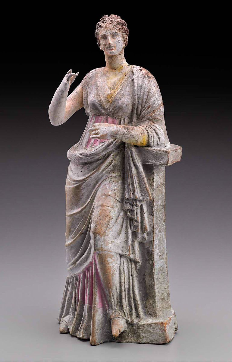 A young woman wearing a high-belted tunic (chiton) and a mantle (himation) leans against a rectangular pillar with a block-like top and places one foot on its base. Her hair is arranged in a "melon-coiffure" or "corn rows", pulled back to a bun at the base of her skull. Loose locks fall from the bun onto her neck. The woman wears a large ring on her ring finger. A pin-hole in the top of the pillar and another on her shoulder indicate that another figure - presumably Eros - orginally was part of the group. Color is still brilliant on some parts of the figure: especially the flesh, the hair, the chiton, which is of intense pink, with a blue border. Missing: left forefinger, and three fingers of right hand. A crack runs down the right side.

MFA Boston (98.893)