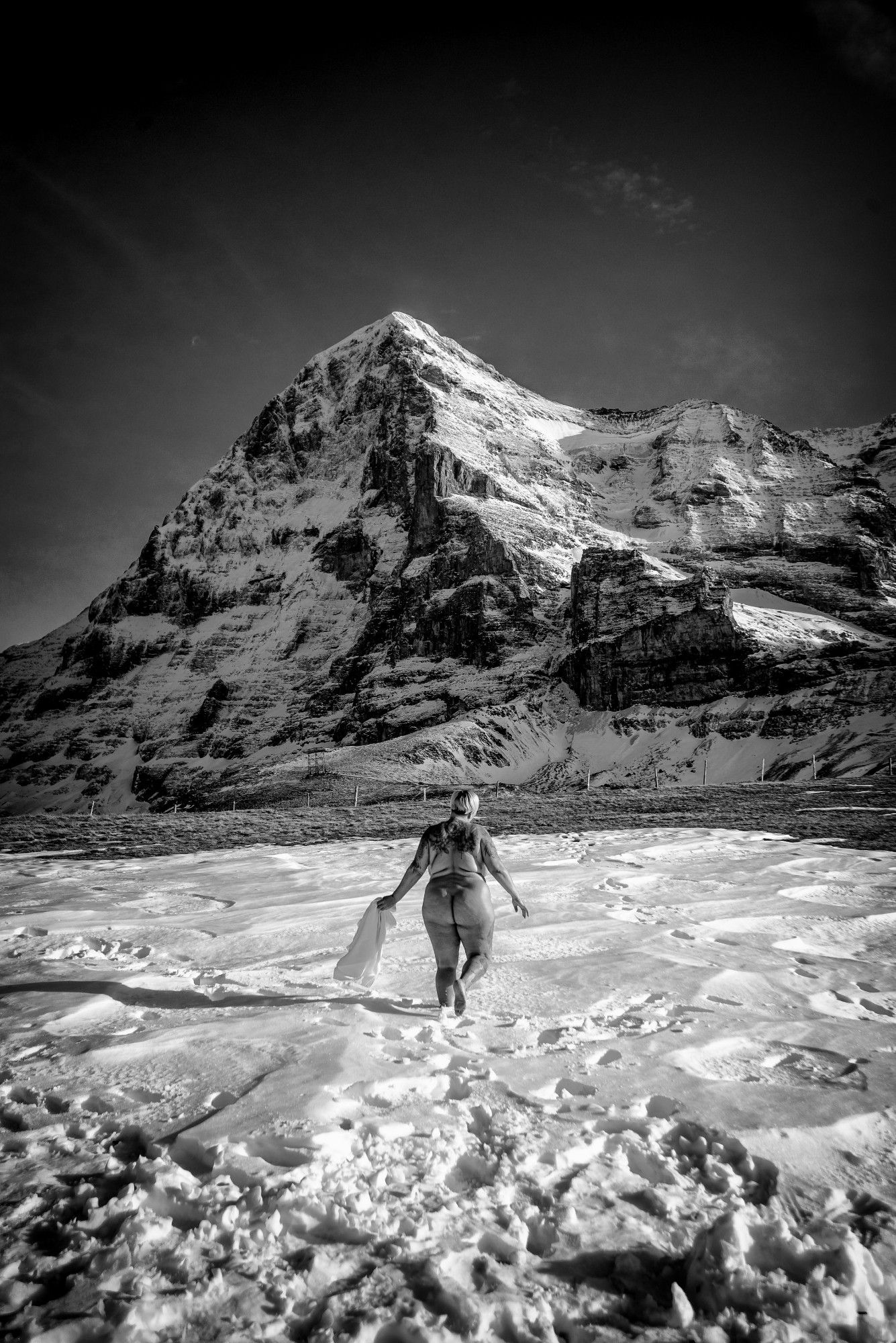 Eine nackte Frau geht durch den Schnee auf die Eiger Nordwand zu. Von hinten gesehen. Schwarzweiß.