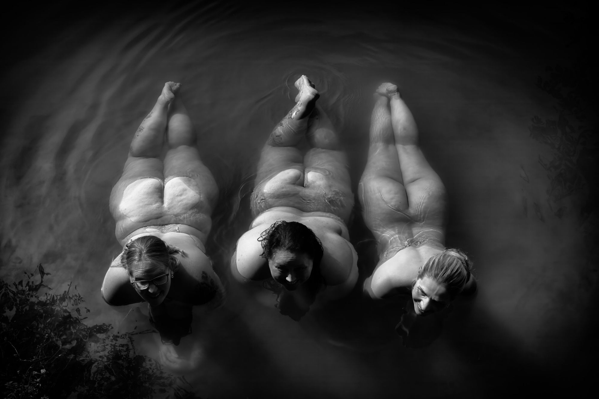 Drei Frauen liegen nebeneinander nackt bäuchlings im flachen Wasser.