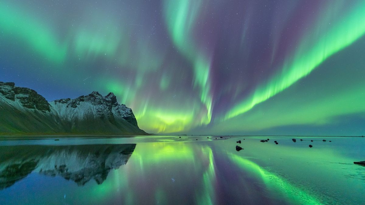 Aurora borealis over water with snow-capped mountains in the background.