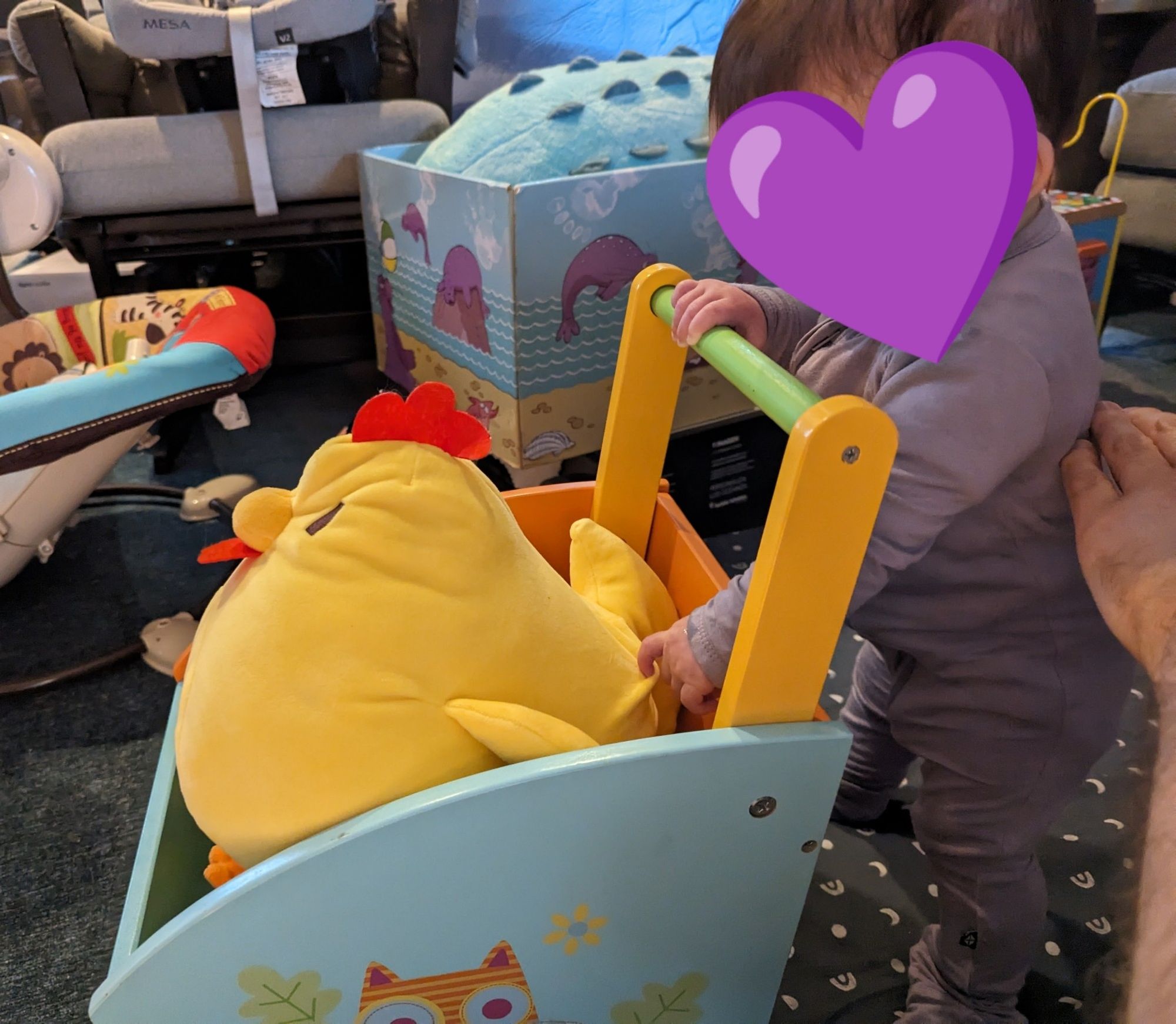 A small baby with dark hair wearing a lavender onesie stands and is pushing a colorful wooden cart. She has one hand on the cart, and one outstretched trying to reach a large yellow chicken plushie in the cart.