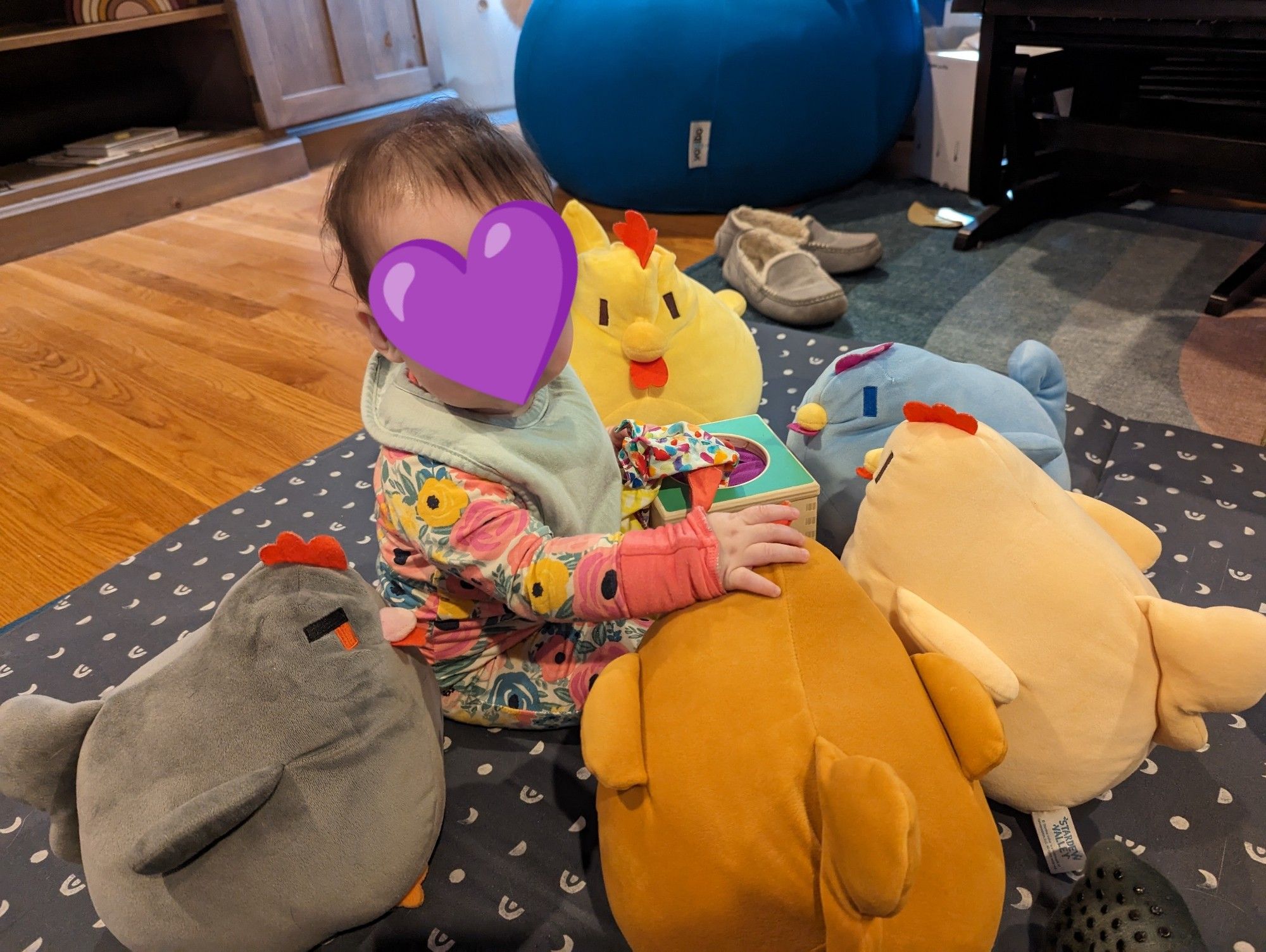 A dark haired baby in a pink floral onesie and a light green bib sits upright surrounded by Stardew Valley chicken plushies. Her face is obscured by a purple heart, and a tissue box toy is in the center with her.

Her real life expression is that of bewildered wonder!