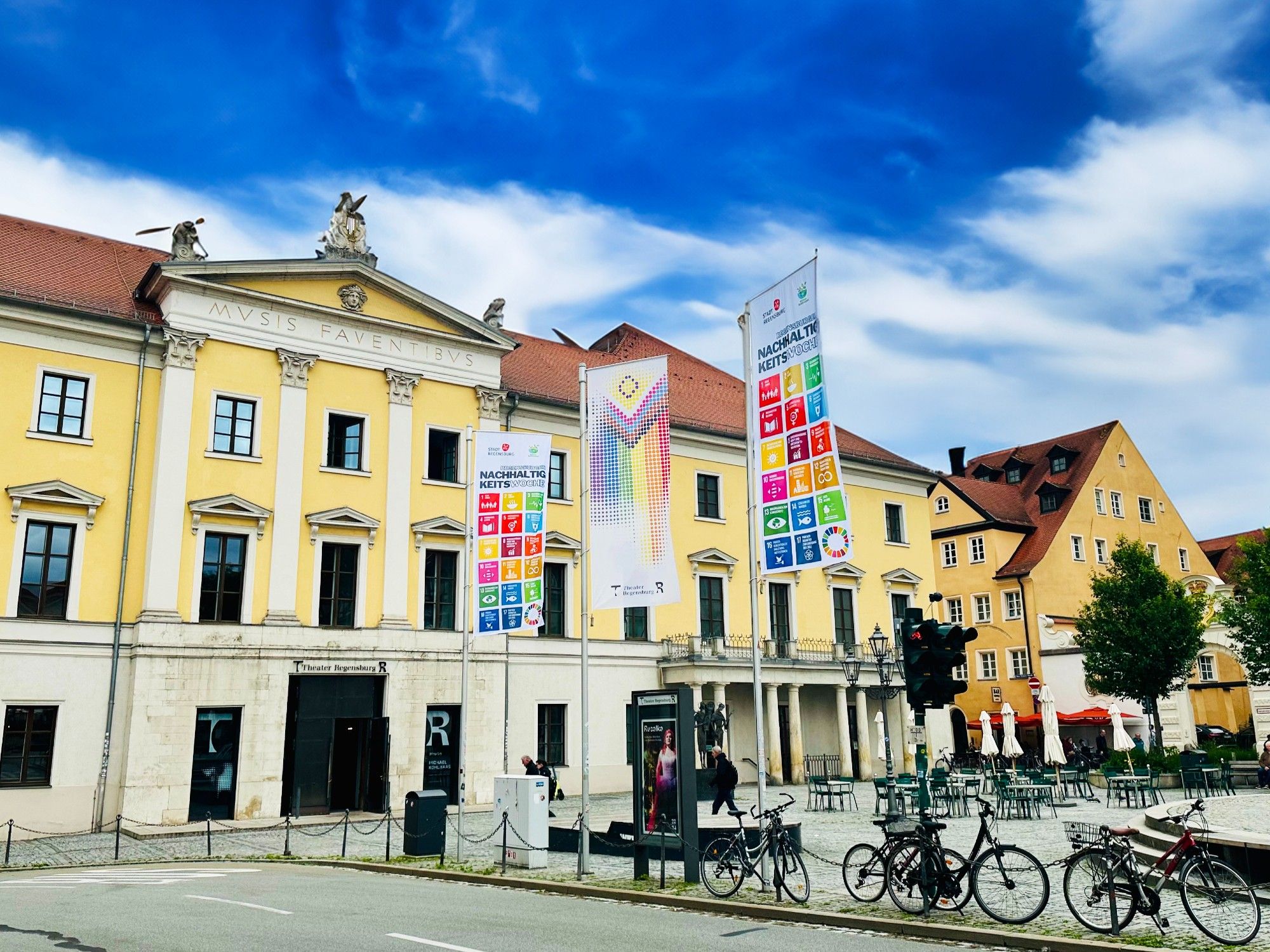 Das gelbe, historische Theatergebäude am Bismarckplatz in Regensburg. Davor zwei Fahnen der Nachhaltigkeitswoche und eine Pride-Flag im Design des Theaters.