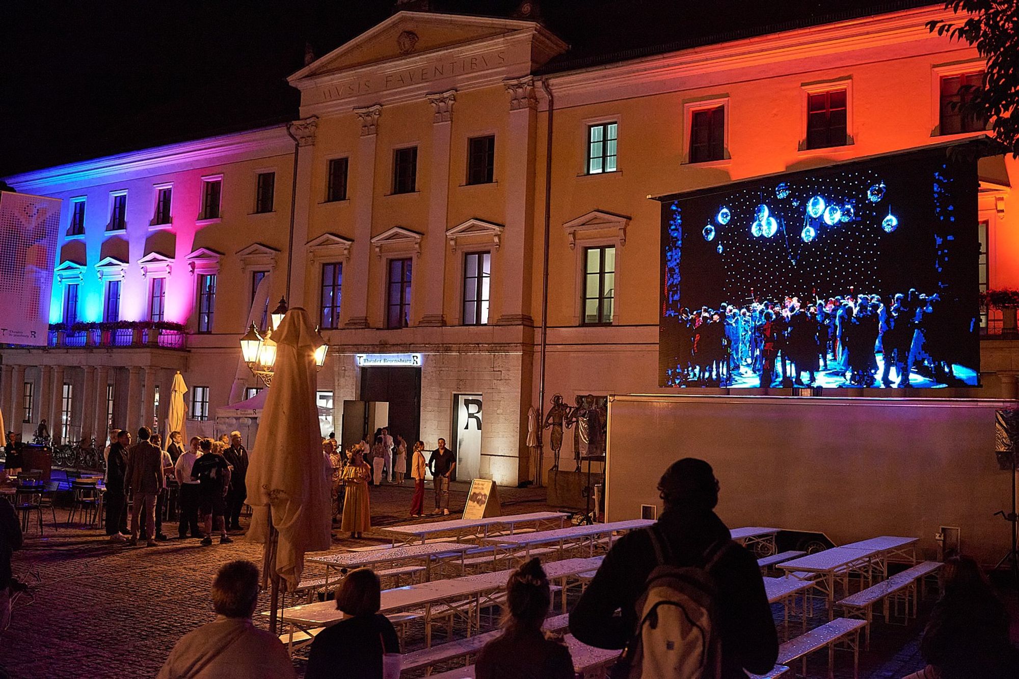 Bunt erleuchtetes Theatergebäude, davor auf der rechten Bildseite eine Großleinwand mit Übertragung aus dem Theatersaal voller tanzender Menschen.