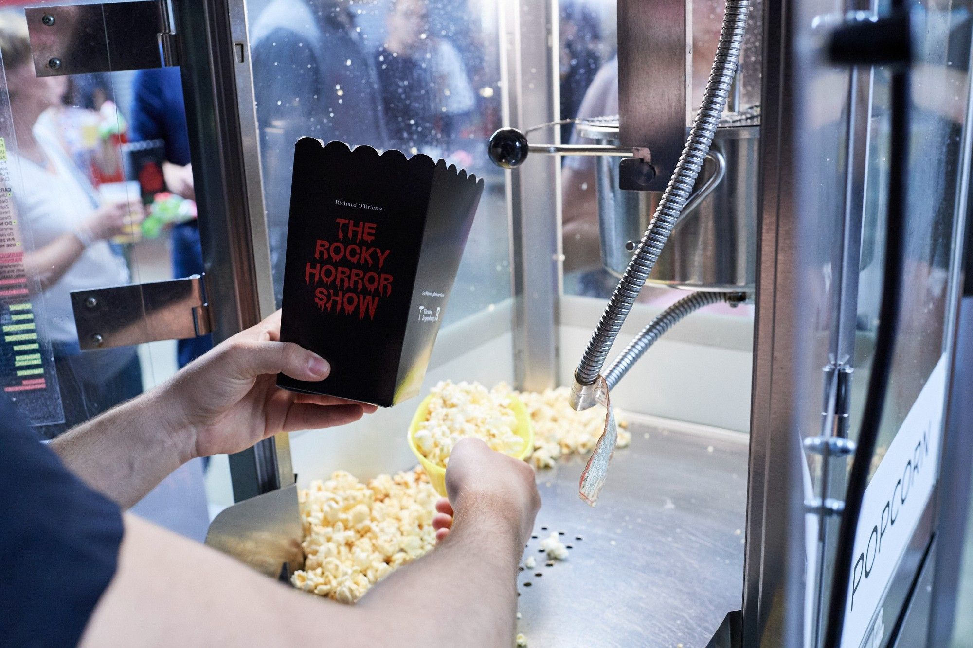 Nahaufnahme einer schwarzen Popcorn-Schachtel mit ROCKY HORRORO SHOW Aufschrift in Rot, in die im Glaskasten einer Popcorn-Maschine gerade Popcorn eingefüllt werden.