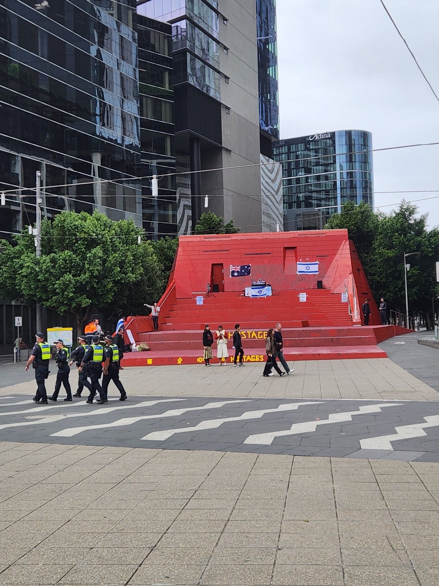 A few people in Melbourne preparing for a rally. Australian and israeli flags are taped on Southbank's red stage. 5 cops walk past.