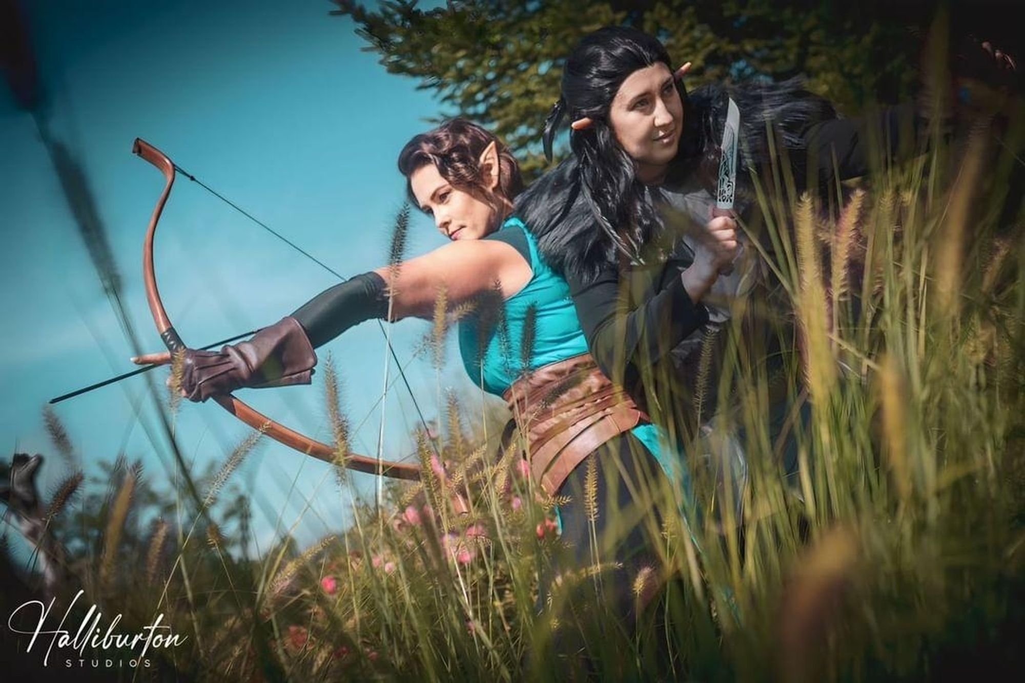 Vexhalia from Legends of Vox Machina is back to back with Vaxildan with her on the left and him on the right. Vex has her bow drawn with an arrow pointed left while Vax has two daggers out and is facing right. They're standing in some tall grass.