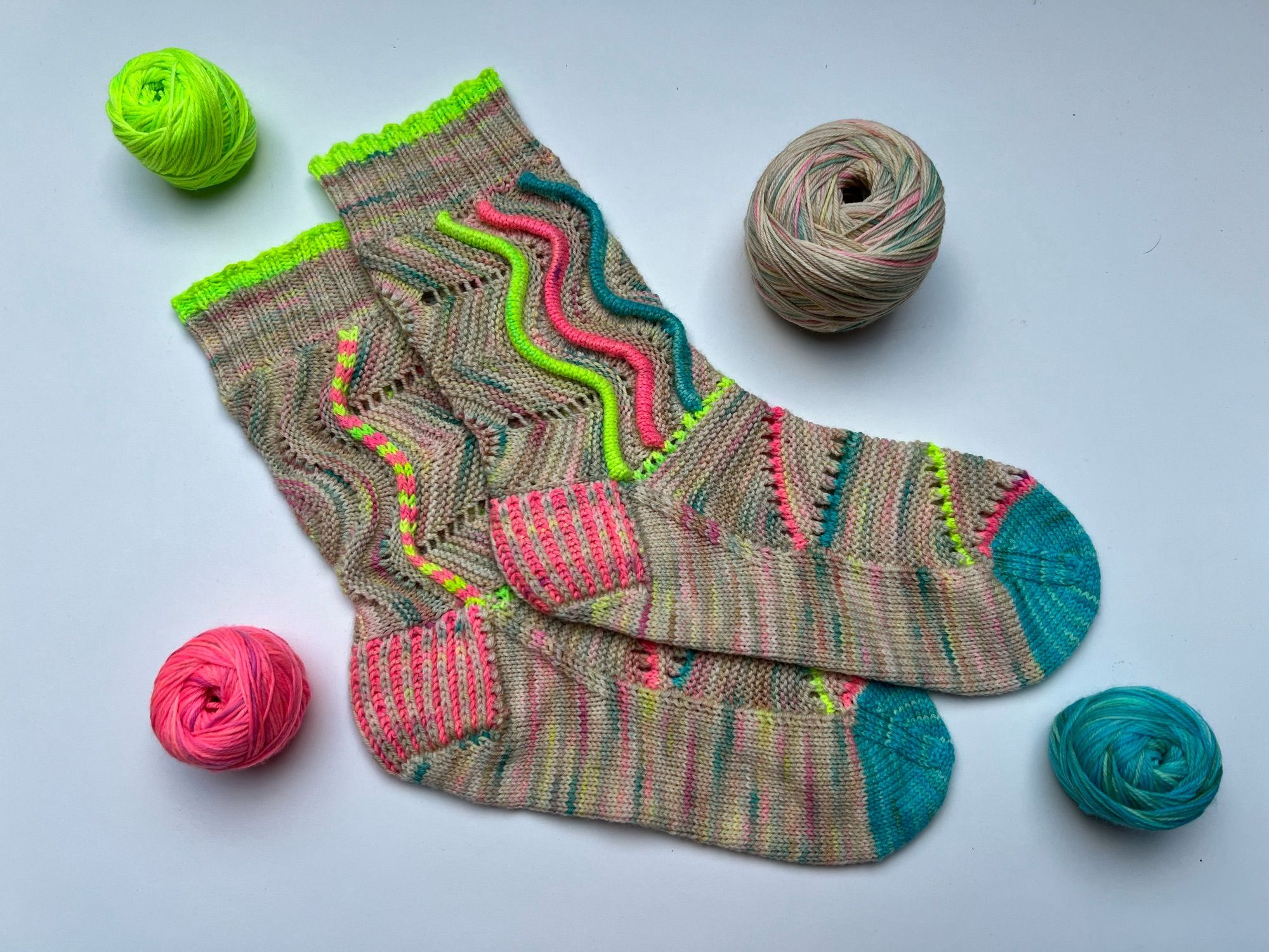 Handknitted pair of socks with a wavy welts pattern, in a neon speckled neutral main colour and matching  neon red, green and blue highlights, shown on a white background with the leftover balls of yarn next to it.