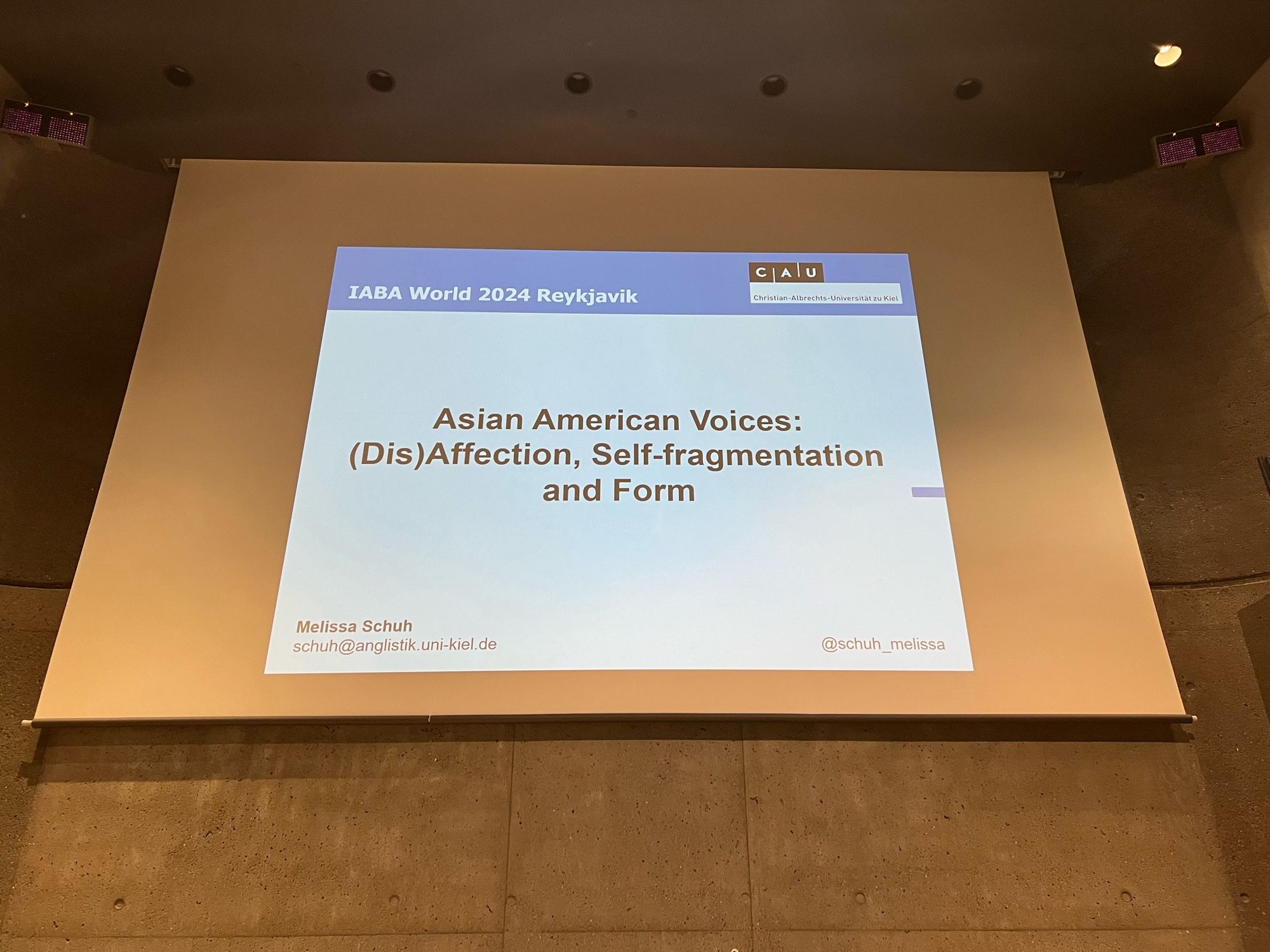 Photo of a power point slide projection on a large screen, showing the title of a talk: Asian American Voices: (Dis)Affection, Self-fragmentation and Form, by Melissa Schuh from the university of Kiel, for the IABA World 2024 Reykjavik conference.