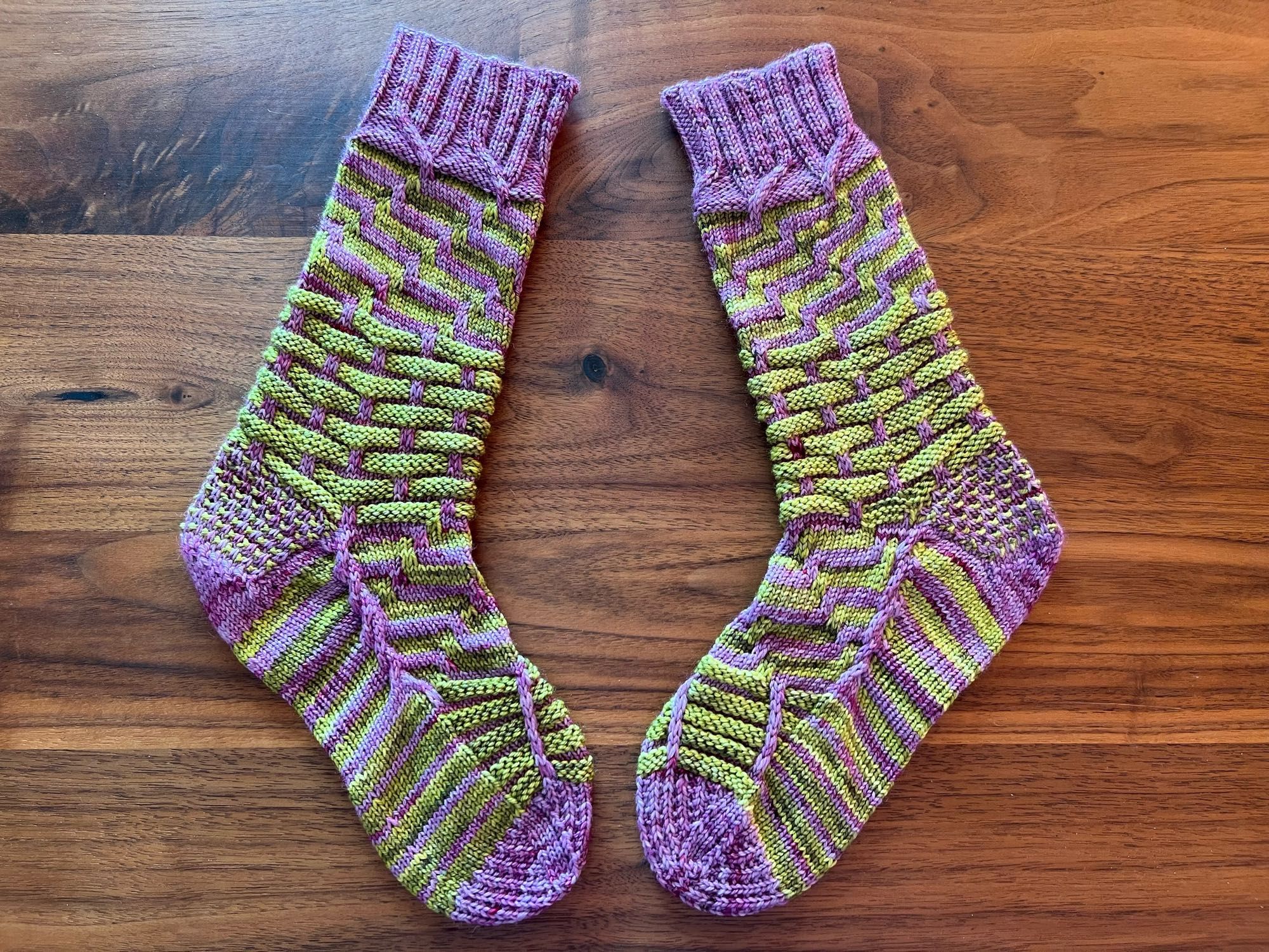 Handknitted pair of socks in a contrasting stripey pattern, knitted in a green and purple semisolid yarn, laid out on a dark wooden table.