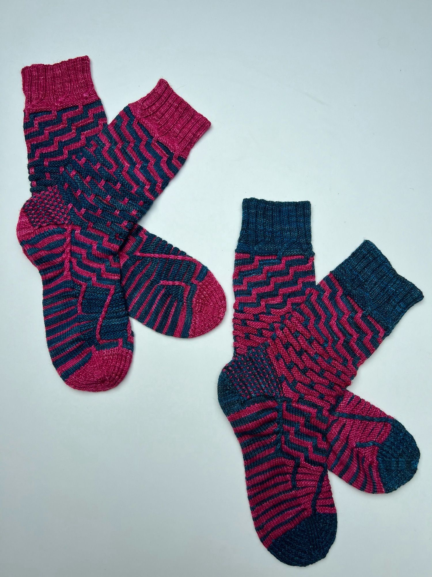 Two handknitted pairs of stripey patterned socks in a contrasting semi-solid teal and magenta yarn, in reverse colour combination, laid out on a white background.