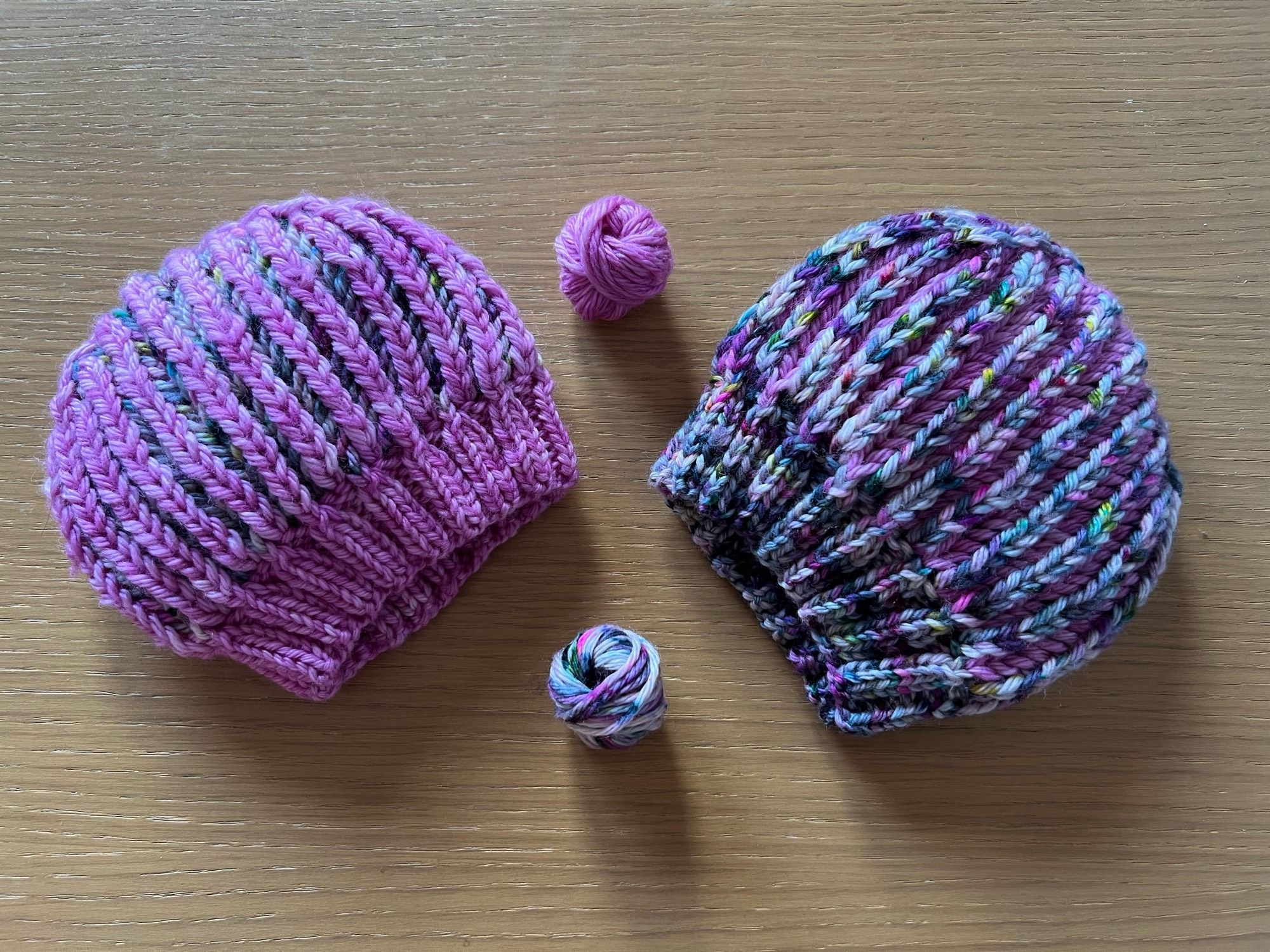 Two Hand knitted reversible Brioche hats, one in a variegated gray, pink, green, light brown yarn with a semi-solid pink contrasting yarn on the inside, and the reverse pink with the variegated inside, laid out on a wooden table.