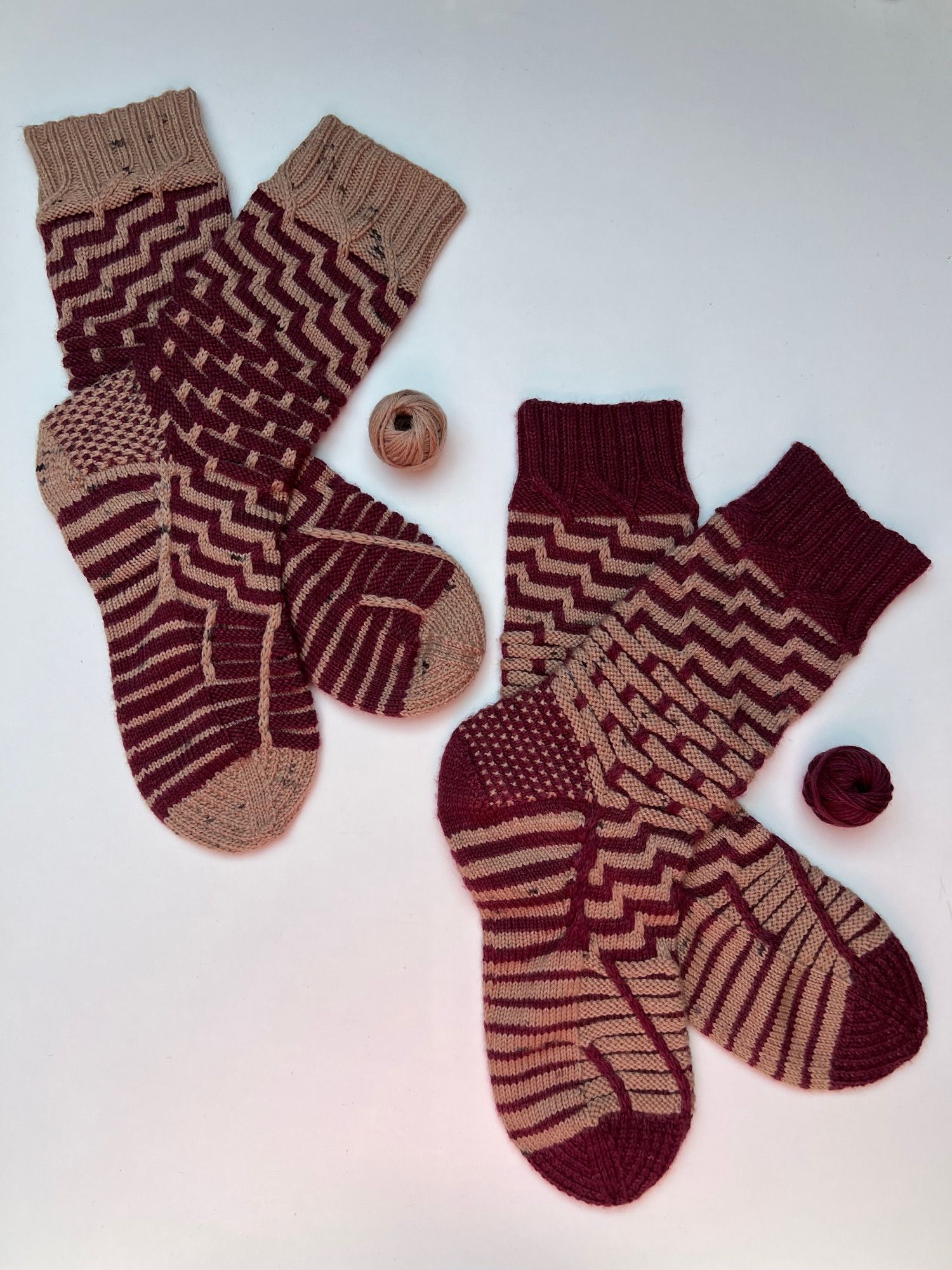 Two pairs of handknitted socks in solid dark red and light brown yarn, in a stripey pattern, one with red as the main colour, the other one in the reverse with brown as the main colour, laid out on a white background.