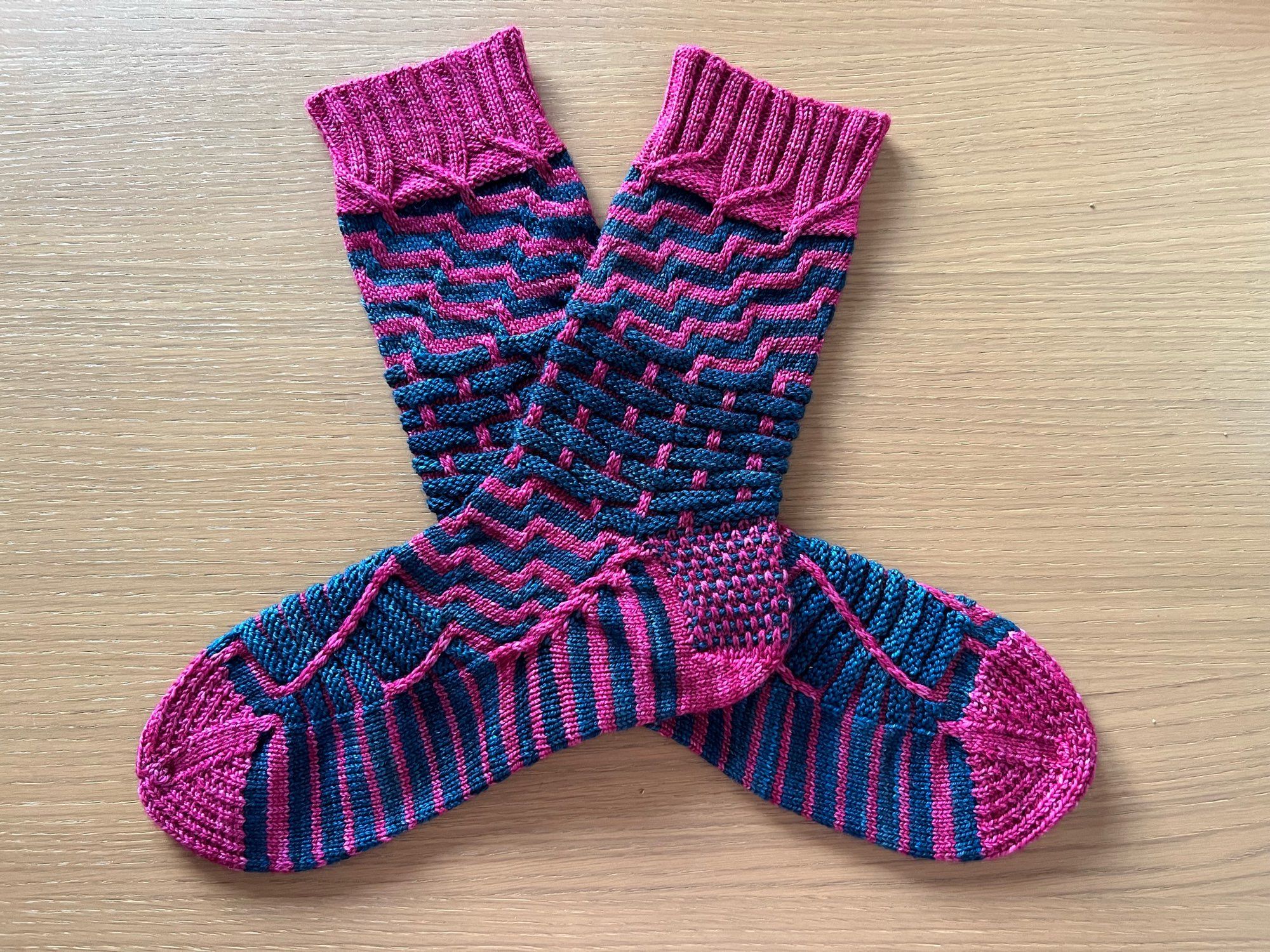 A pair of hand knitted  pairs of socks in a semi-solid dark teal and magenta pink yarn in a complex pattern of contrasting stripes and textured, laid out on a wooden table.