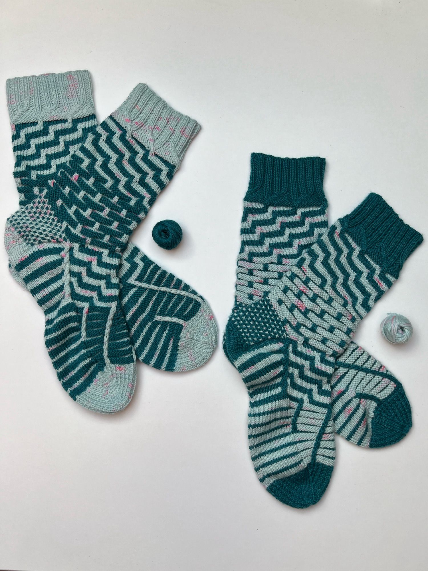 Two pairs of handknitted socks in solid dark teal and aqua yarn, in a stripey pattern, one with teal as the main colour, the other one in the reverse with aqua as the main colour, laid out on a white background.