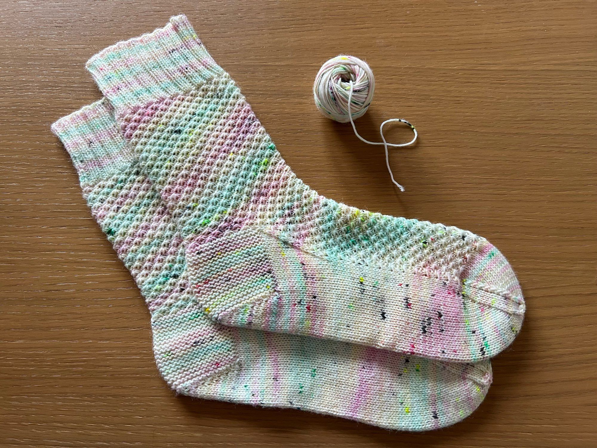 Handknitted pair of socks in a textured pattern,  in a variegated pastel pink, blue, green, and cream yarn, laid out on a wooden table with the leftover ball of yarn next to it.