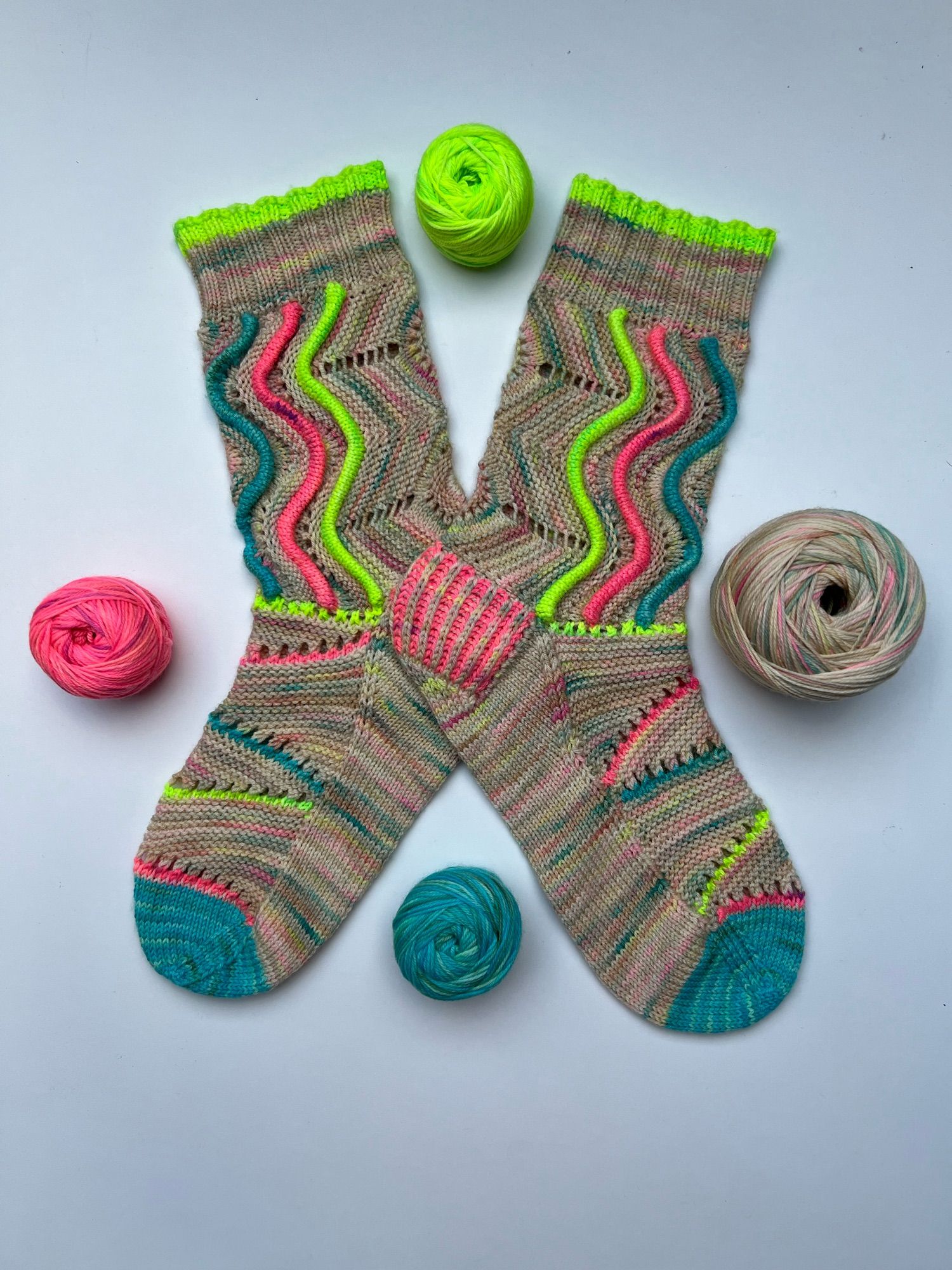 Handknitted pair of socks with a wavy welts pattern, in a neon speckled neutral main colour and matching  neon red, green and blue highlights, shown on a white background with the leftover balls of yarn next to it.