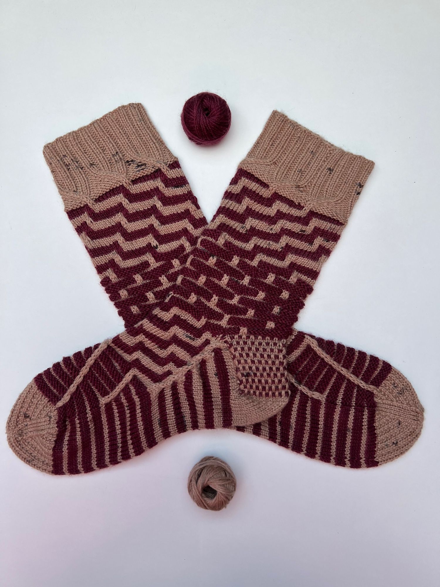 One pair of handknitted socks in solid dark red and light brown yarn, in a stripey pattern, with brown as the main colour, laid out on a white background with the tiny balls of leftover yarn next to it.