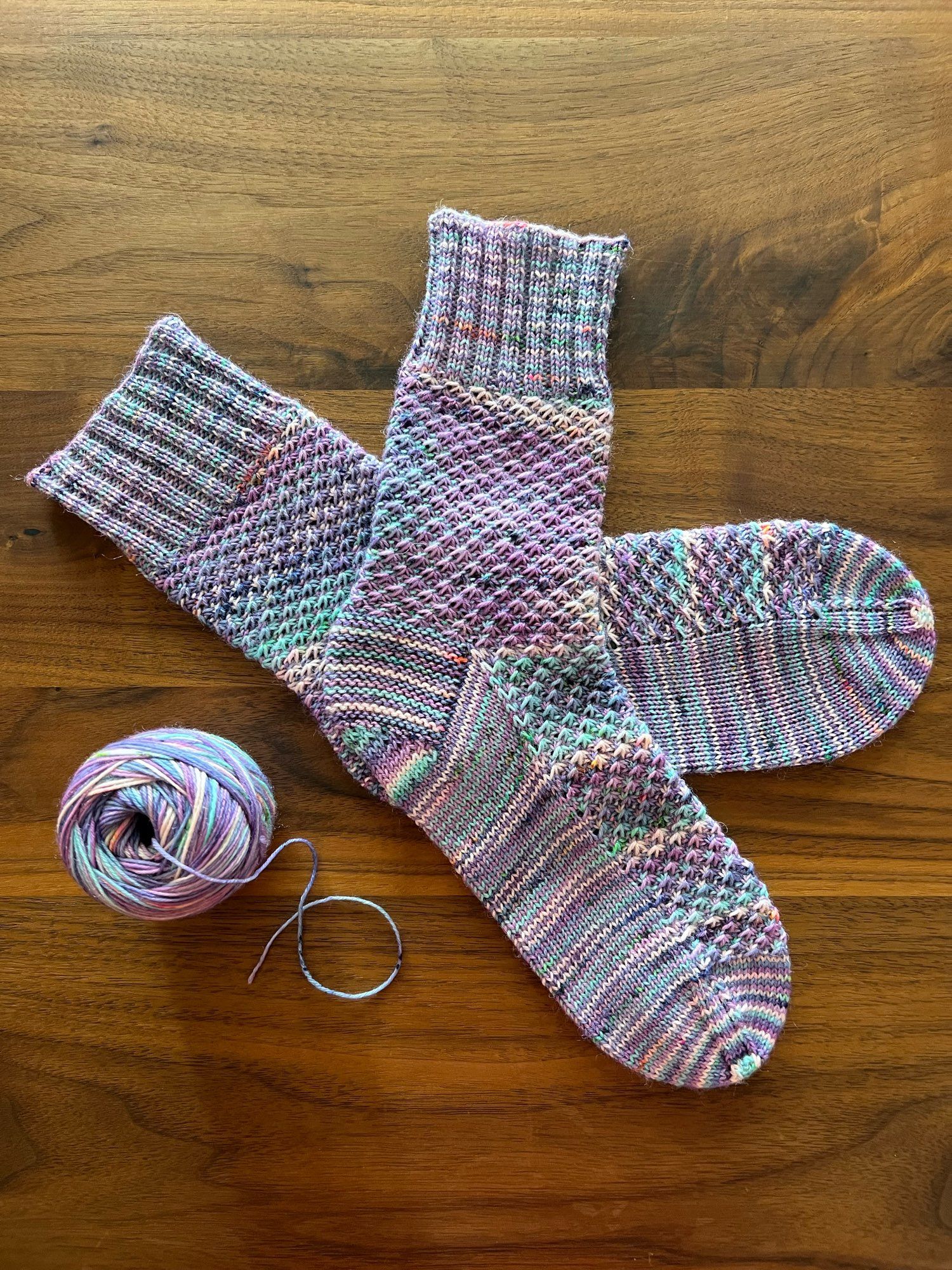 Handknitted pair of socks in a textured pattern, knitted in a variegated purple, peach, blue and light green yarn with orange speckles, laid out in a wooden table with the leftover ball of yarn next to it.