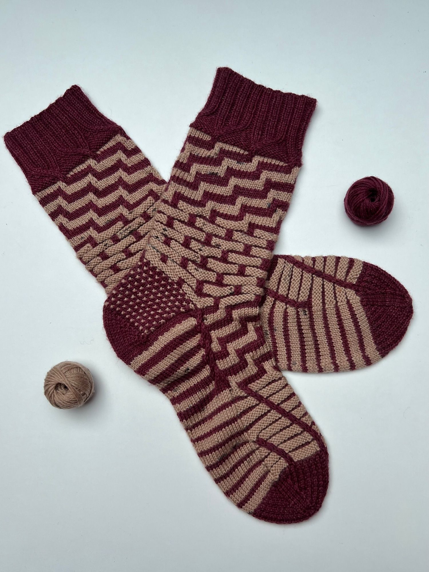 One pair of handknitted socks in solid dark red and light brown yarn, in a stripey pattern, with red as the main colour, laid out on a white background with the tiny balls of leftover yarn next to it.