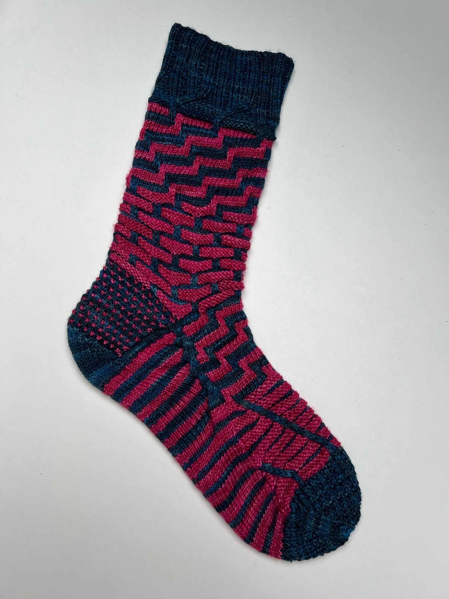 Handknitted sock in a stripey pattern in two contrasting semi solid hand dyed yarn colours, a dark teal and a bright magenta pink, shown on a white background.