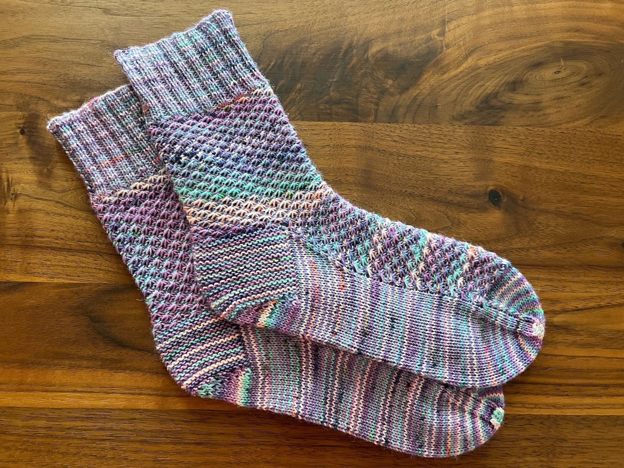 Handknitted pair of socks in a textured pattern, knitted in a variegated purple, peach, blue and light green yarn with orange speckles, laid out in a wooden table.