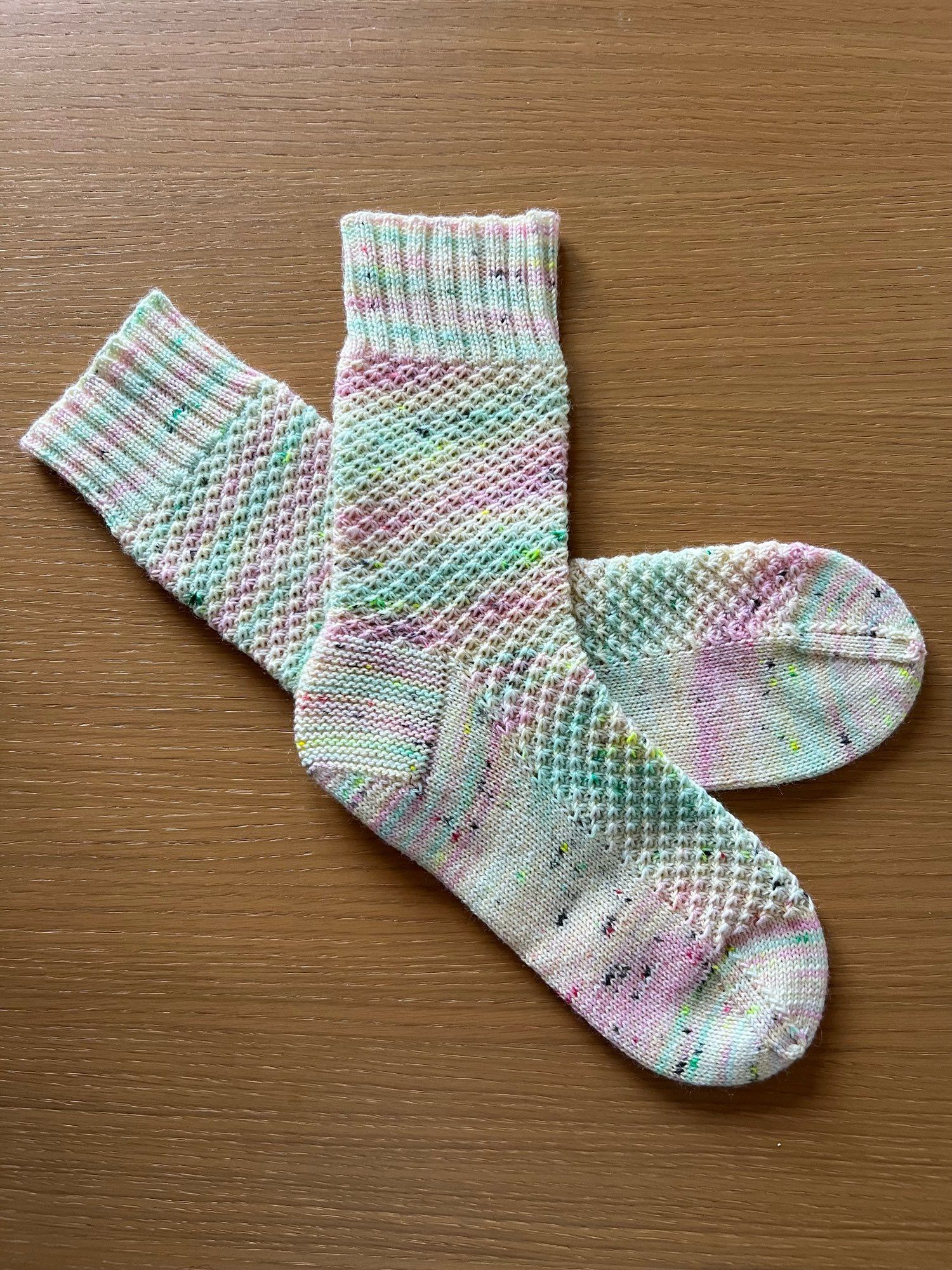 Handknitted pair of socks in a textured pattern,  in a variegated pastel pink, blue, green, and cream yarn.