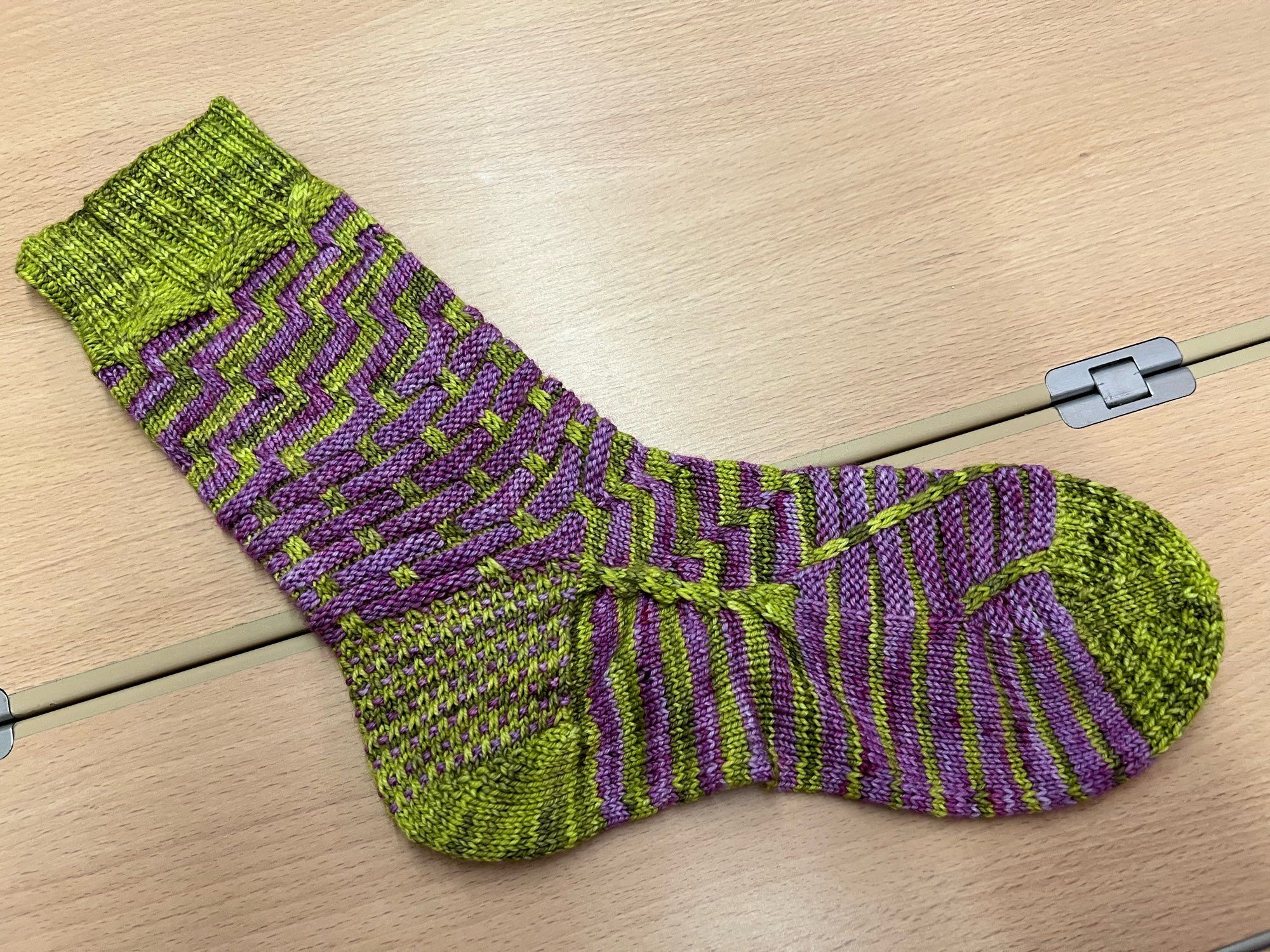 Handknitted sock in a contrasting stripey pattern, knitted in a green and purple semisolid yarn, laid out on a wooden table.