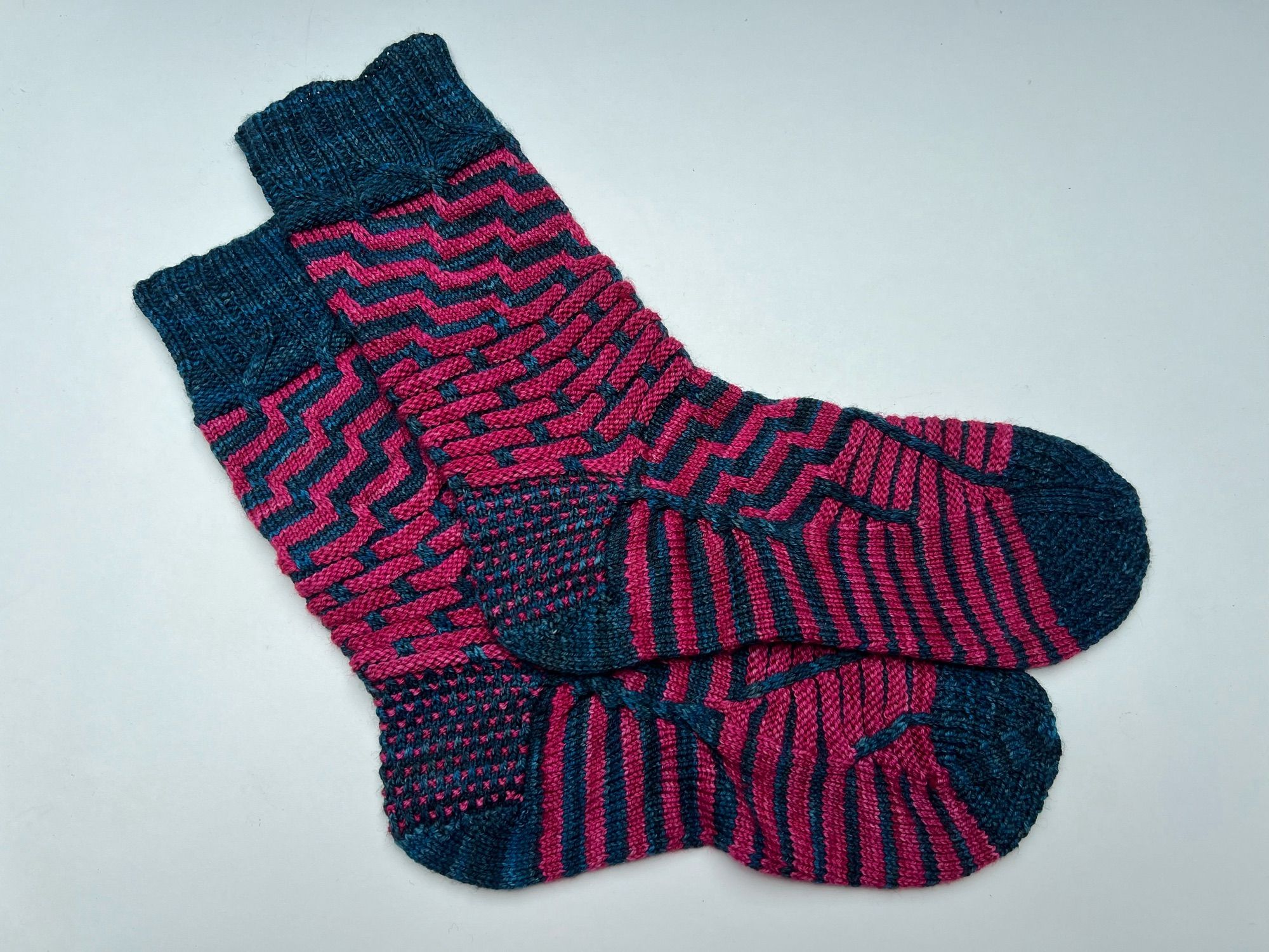 Handknitted pair of stripey patterned socks in a contrasting semi-solid teal and magenta yarn, laid out on a white background.