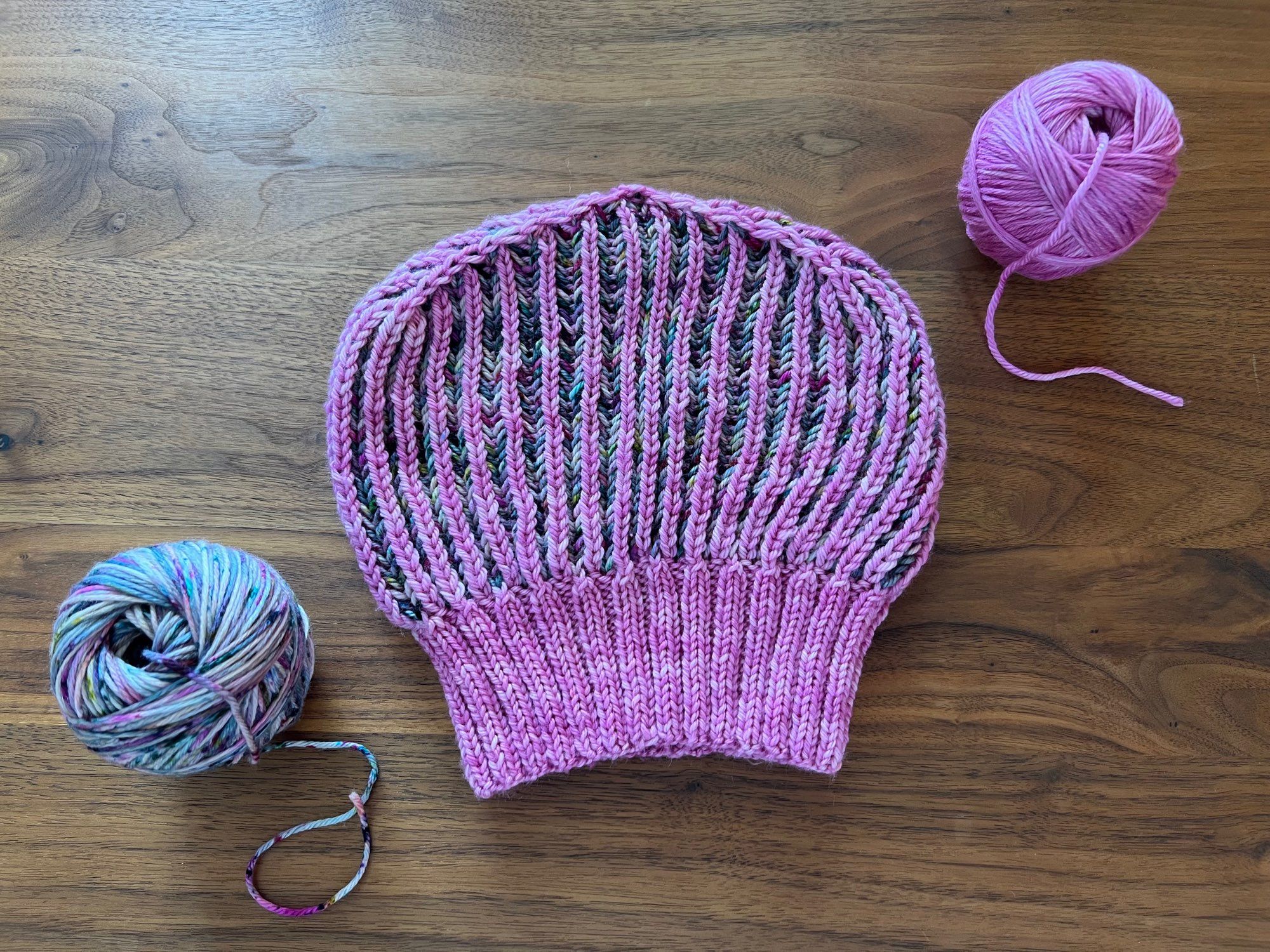 Reversible brioche hat in a semi-solid pink and a variegated magenta, green, grey, and black speckles yarn, laid out on a wooden table with the leftover balls of yarn next to it.