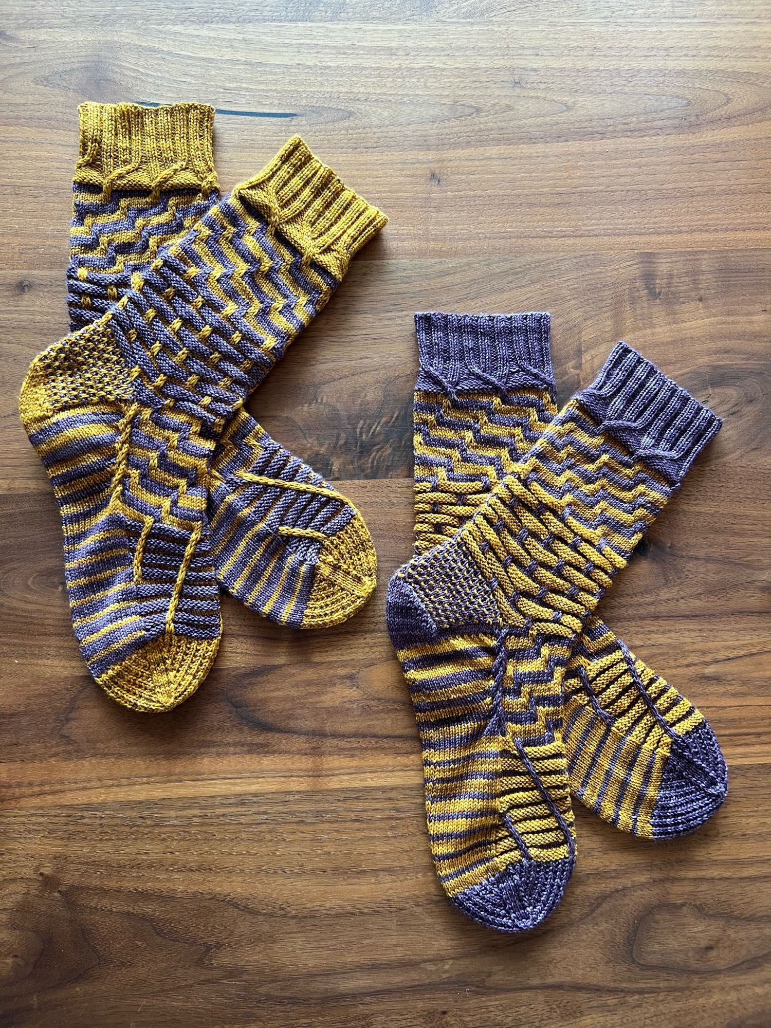 Two reverse coloured hand knitted  pairs of socks in a semi-solid dusky purple and mustard yellow yarn in a complex pattern of contrasting stripes and textured, laid out on a wooden table.