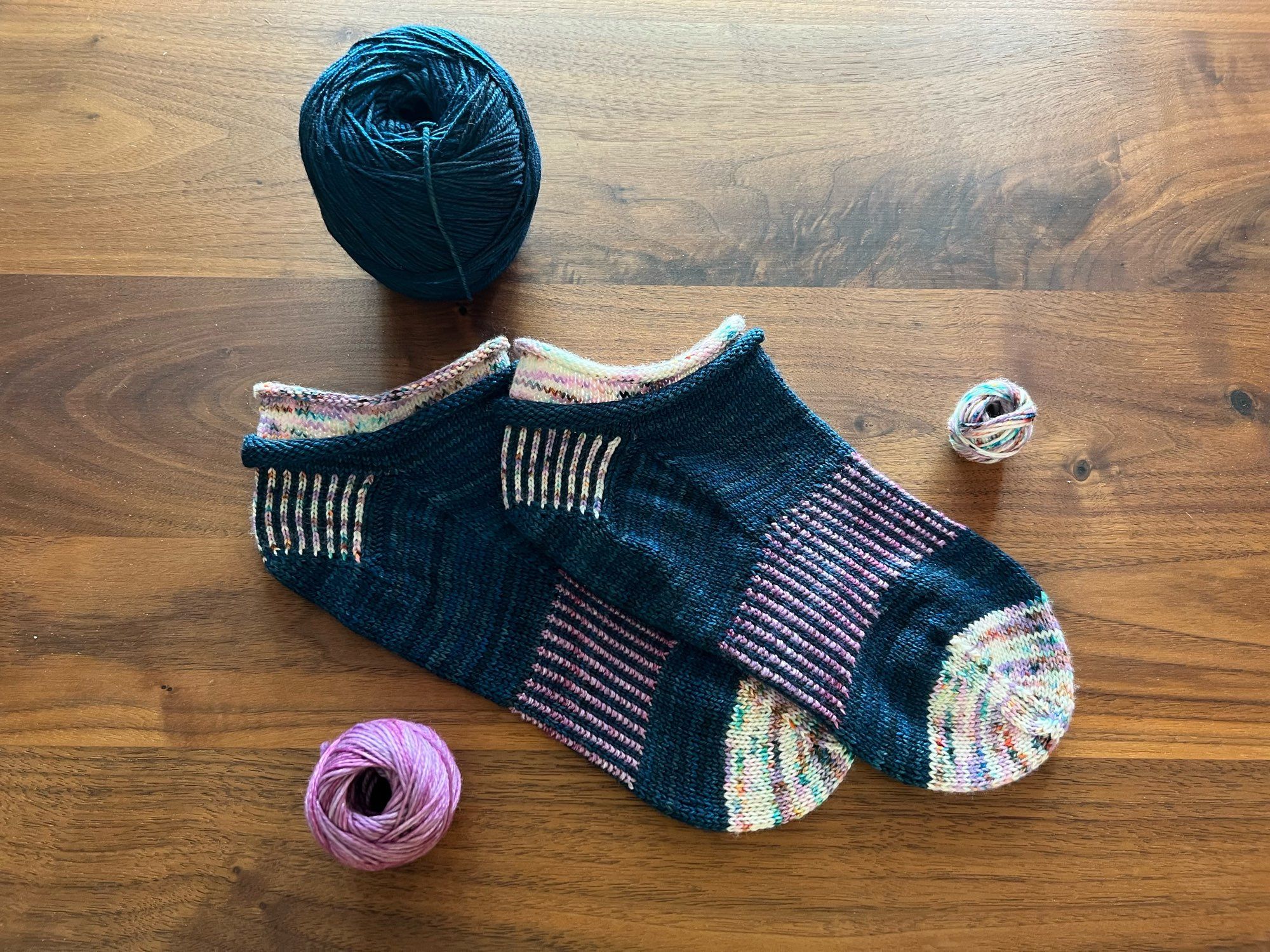 Handknitted Jelly roll ankle socks in a dark teal, purple speckled white, and purple yarn with a contrasting cuff, heel and arch ribbing and toe, laid out on a dark wooden table.