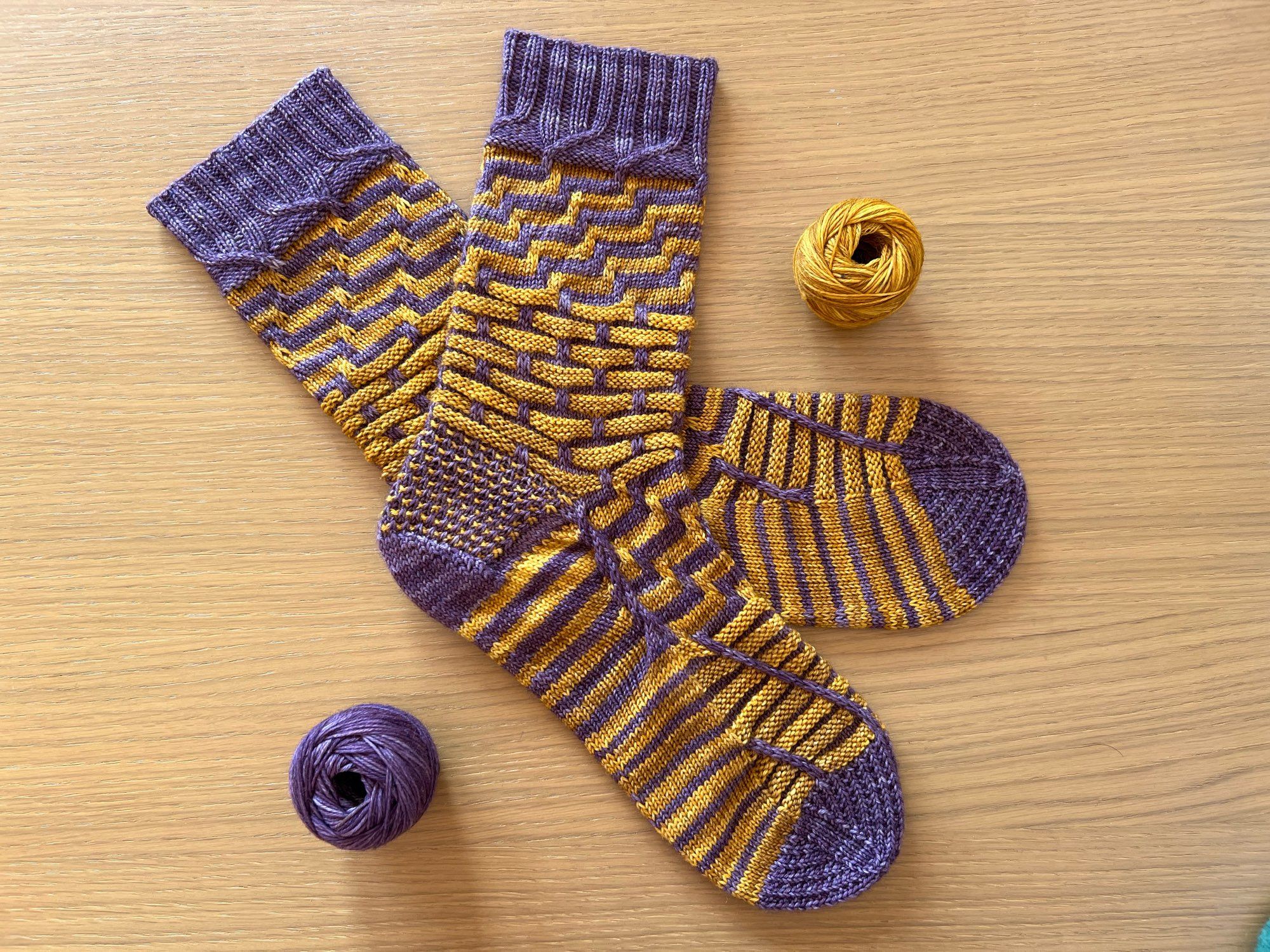 Handknitted pair of socks in a semi-solid dusky purple and mustard yellow yarn in a complex pattern of contrasting stripes and textured, laid out on a wooden table with the leftover balls of yarn next to it.