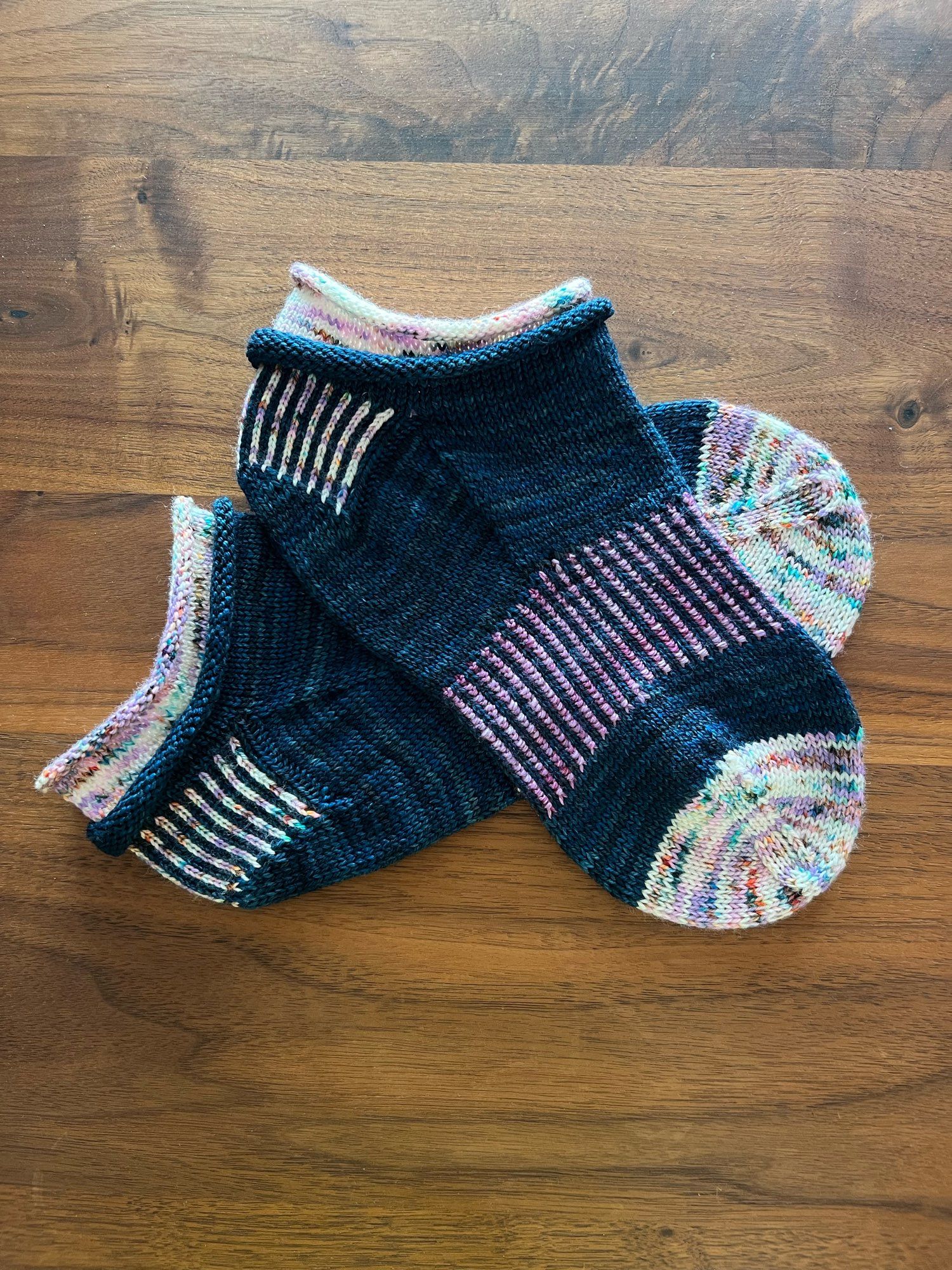 Handknitted Jelly roll ankle socks in a dark teal, purple speckled white, and purple yarn with a contrasting cuff, heel and arch ribbing and toe, laid out on a dark wooden table.