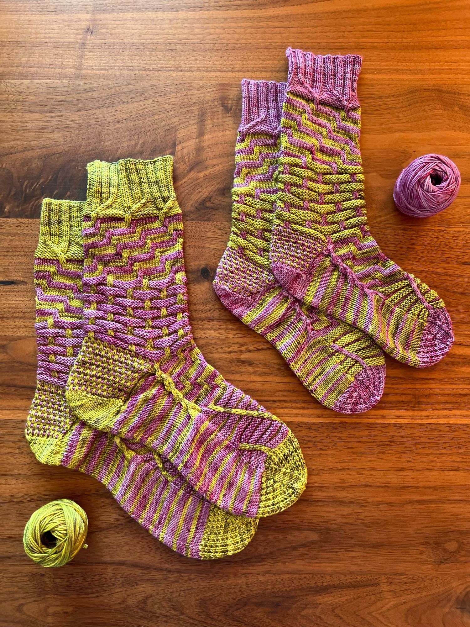 Two hand knitted pairs of socks in reverse colour combination in a contrasting stripey pattern, knitted in a green and purple semisolid yarn, laid out on a dark wooden table with the leftover balls of yarn next to it.