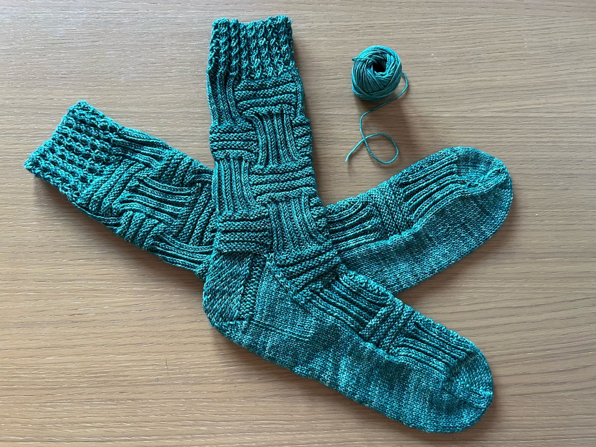 Hand knitted pair of socks, in a basket weave pattern, in a deep green coloured yarn, laid out on a wooden table with the leftover ball of yarn to one side.