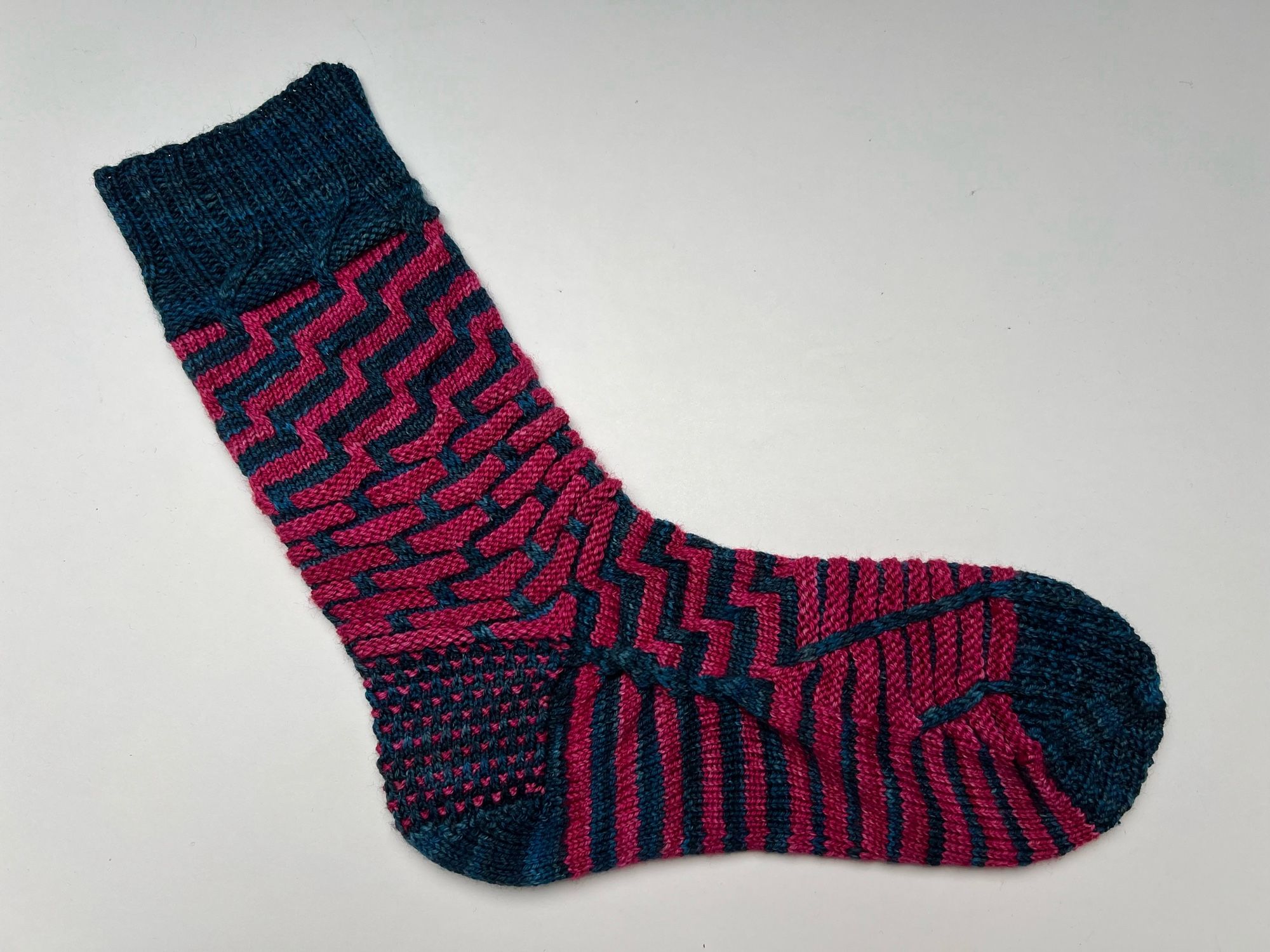 Handknitted sock in a stripey pattern in two contrasting semi solid hand dyed yarn colours, a dark teal and a bright magenta pink, shown on a white background.