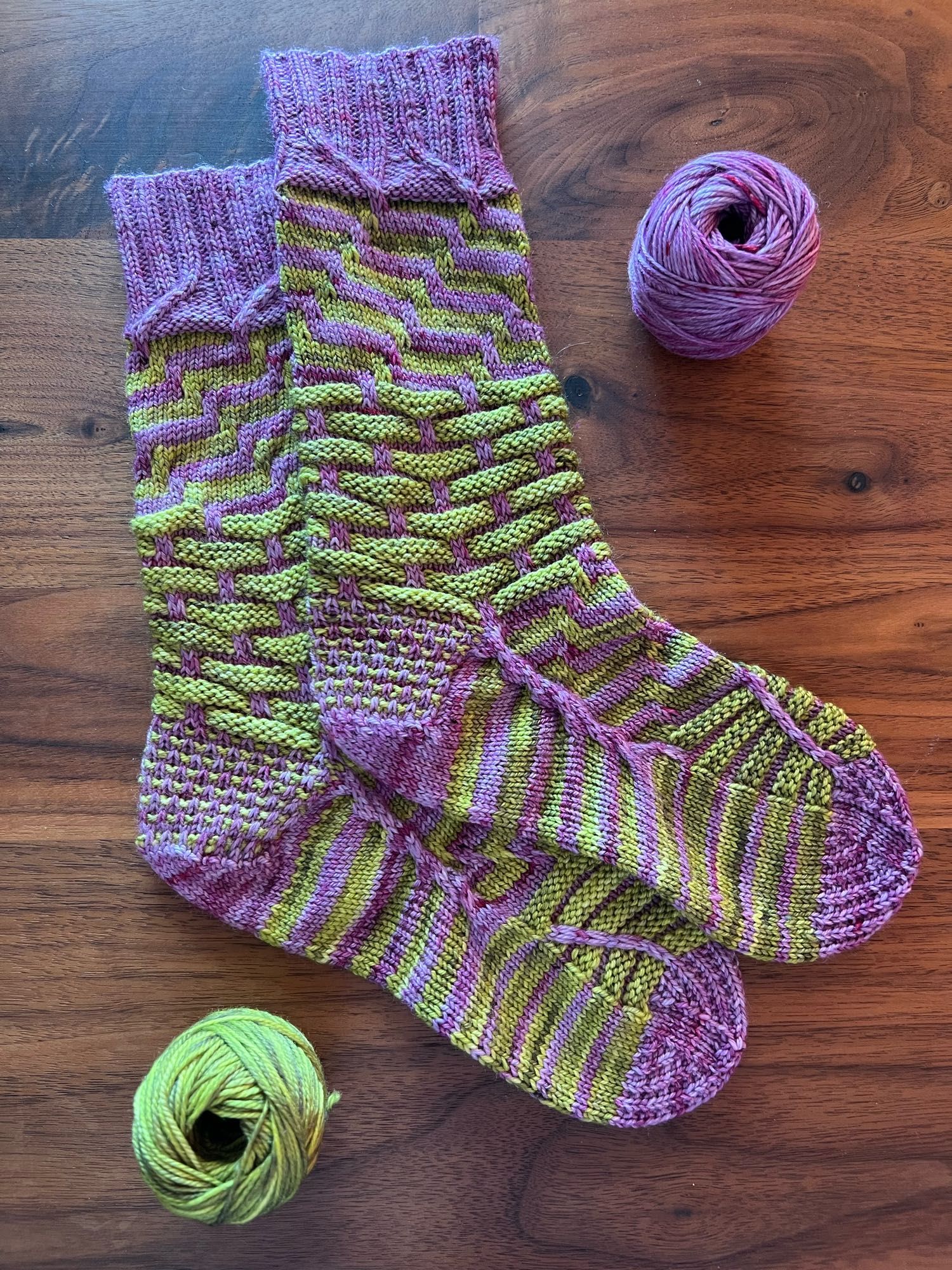 Handknitted pair of socks in a contrasting stripey pattern, knitted in a green and purple semisolid yarn, laid out on a dark wooden table with the leftover balls of yarn next to it.