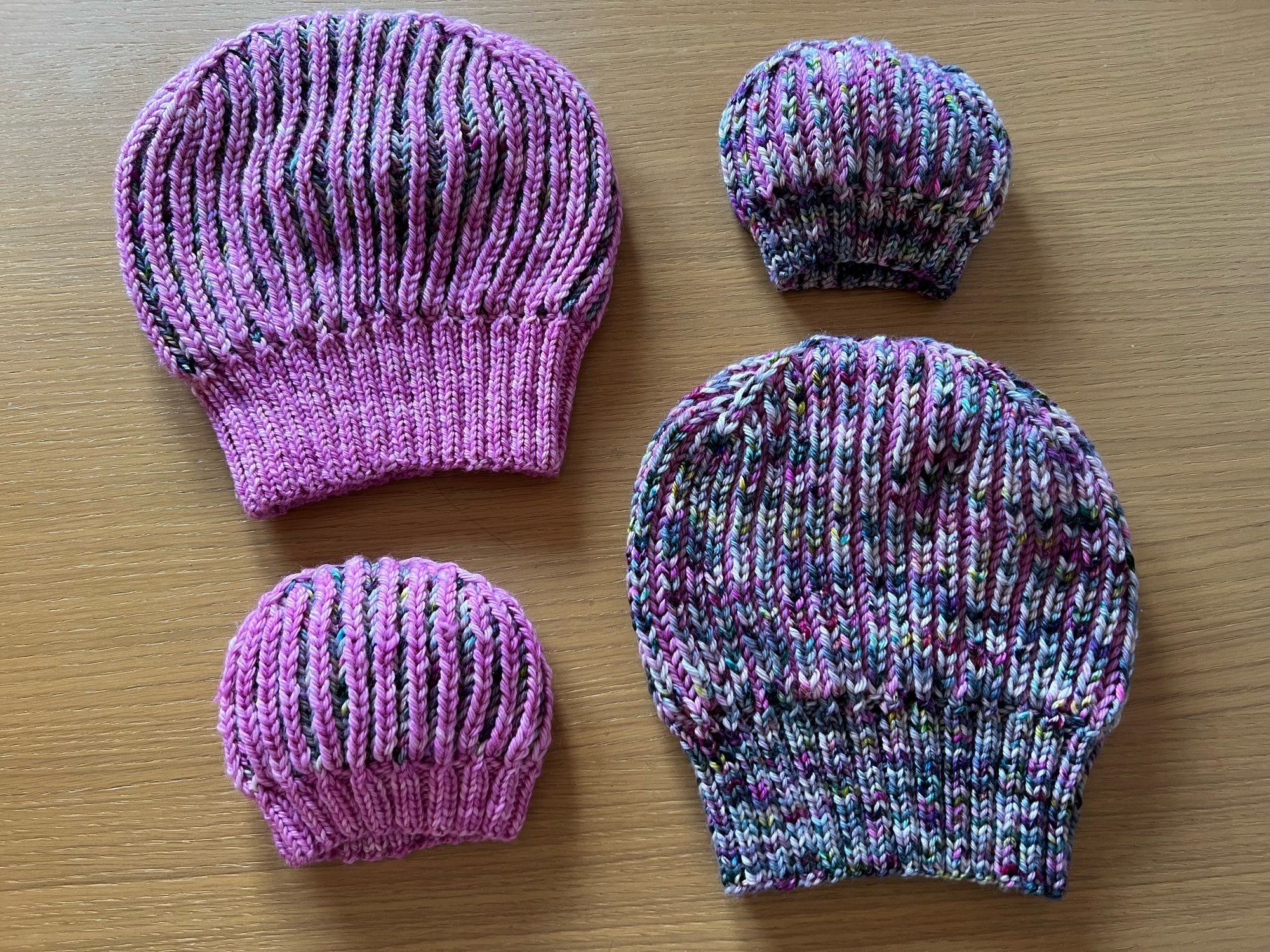 Four Hand knitted reversible Brioche hats (two adult and two baby sizes), one in a variegated gray, pink, green, light brown yarn with a semi-solid pink contrasting yarn on the inside, and the reverse pink with the variegated inside, laid out on a wooden table.