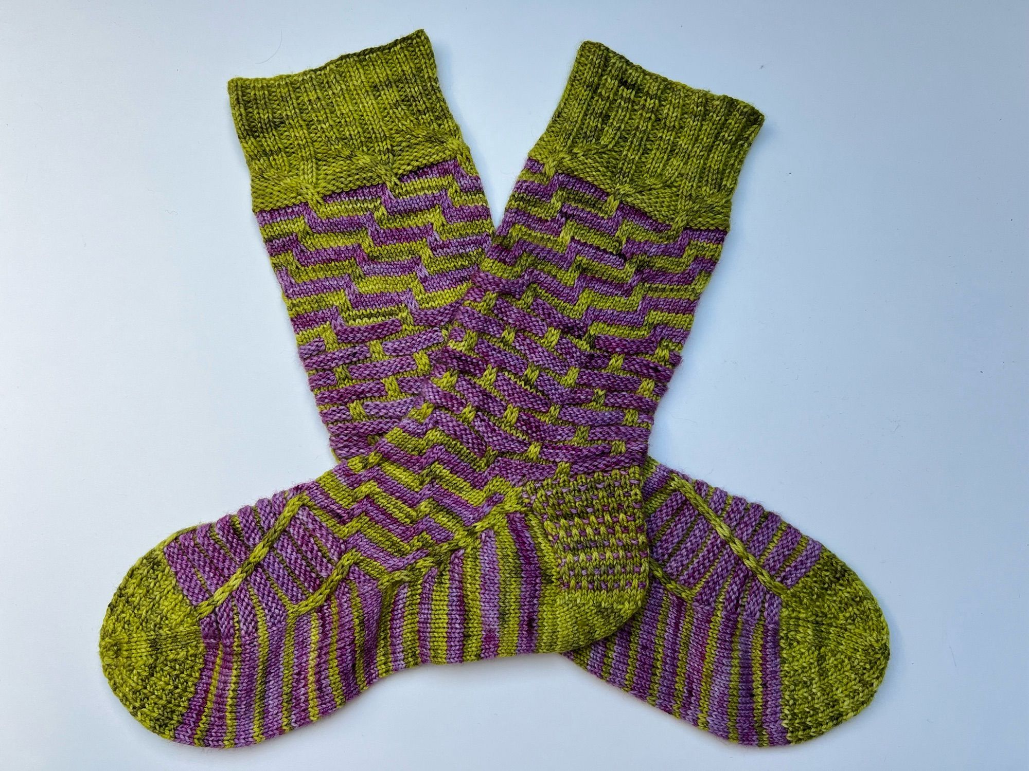 Handknitted pair of socks in a contrasting stripey pattern, knitted in a green and purple semisolid yarn, laid out on a white table.