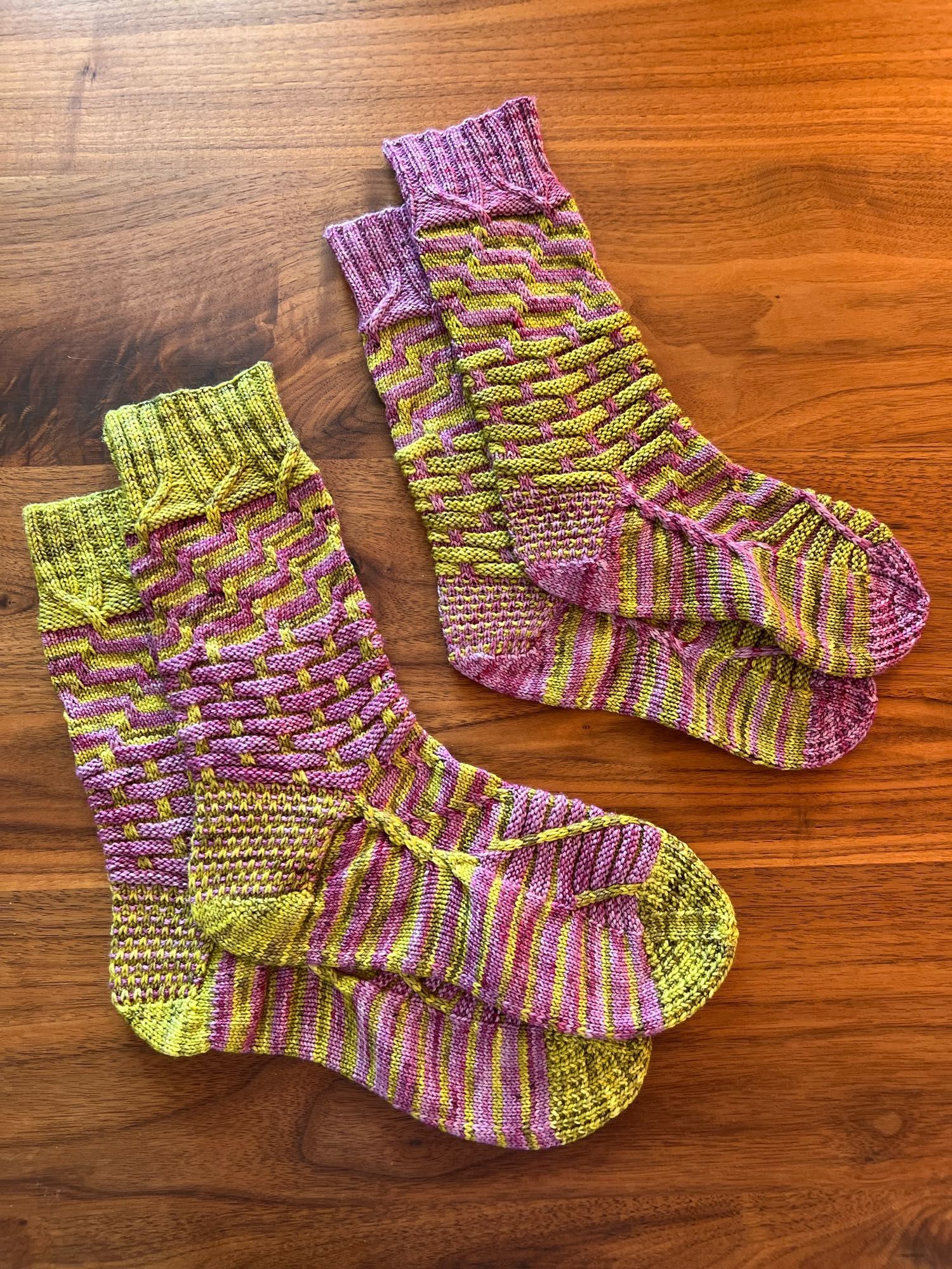 Two hand knitted pairs of socks in reverse colour combination in a contrasting stripey pattern, knitted in a green and purple semisolid yarn, laid out on a dark wooden table.