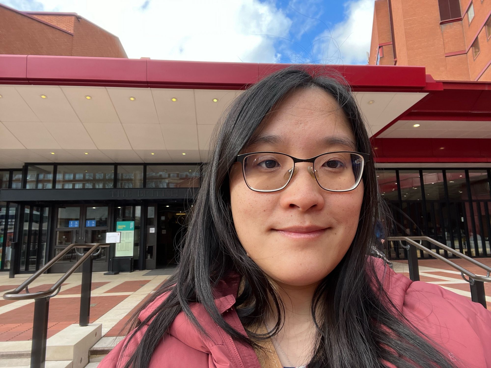 Selfie showing myself, Melissa, an Asian woman with long black hair, wearing a dark pink puffy jacket and glasses, in front of the British Library with the library square and the main building in the background.