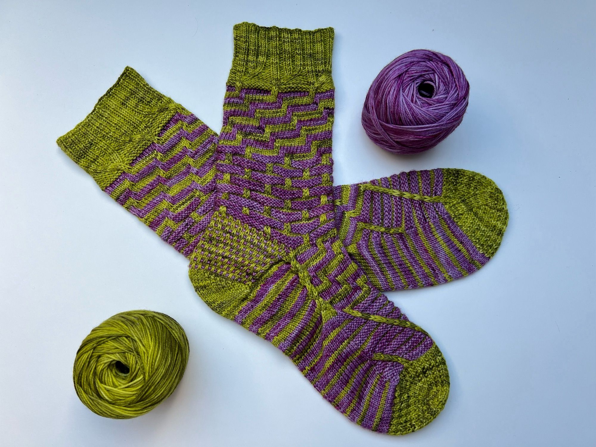 Handknitted pair of socks in a contrasting stripey pattern, knitted in a green and purple semisolid yarn, laid out on a white table with the leftover balls of yarn next to it.