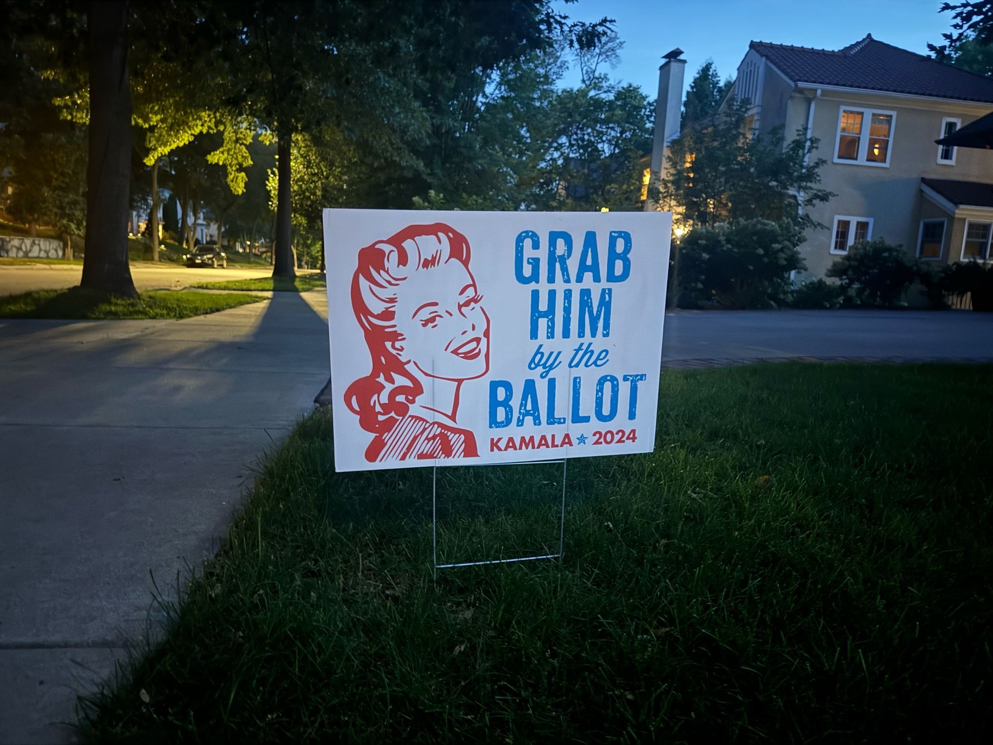 A gorgeous letter-press-style sign the Grab him by the ballot in blue writing next to a drawing of a laughing woman with a roughly 1940s hairstyle. It says Kamala 2024 at the bottom