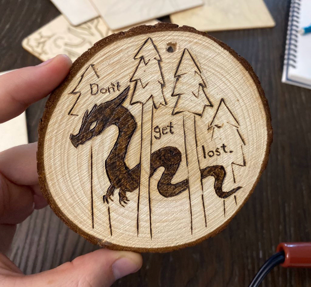 A wooden slab pyrography artwork, small oak circle. It has a design of a dark serpent in some pine trees with the words: “Don’t get lost.”