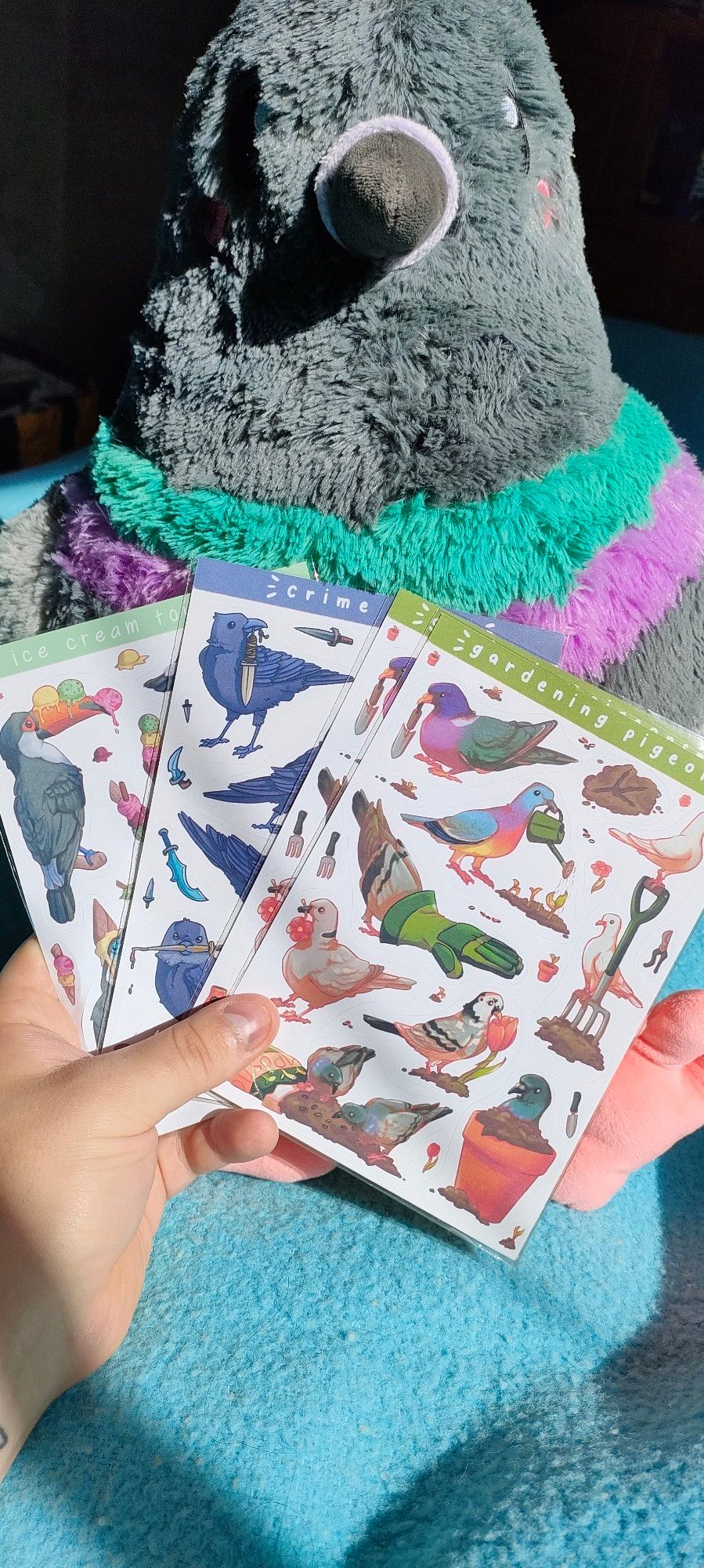 Several pages of stickers held in hand in front of a large pigeon plushie. The stickers display cutesy pigeons in a gardening theme, crows with various weapons, and toucans with ice cream.